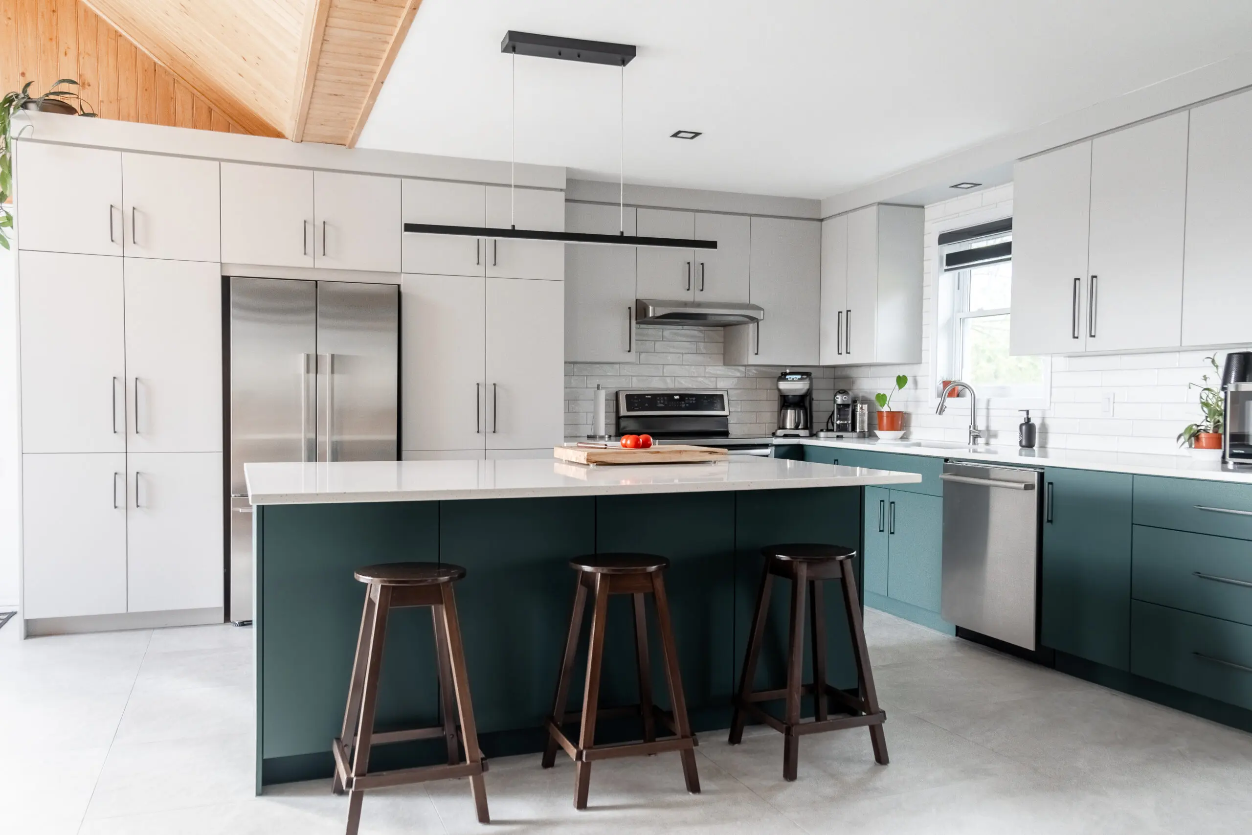 Cuisine moderne avec des armoires blanches et sarcelles, des appareils électroménagers en acier inoxydable, un îlot avec trois tabourets en bois foncé, une coupe de fruits et une élégante lampe suspendue noire au-dessus de l'îlot.