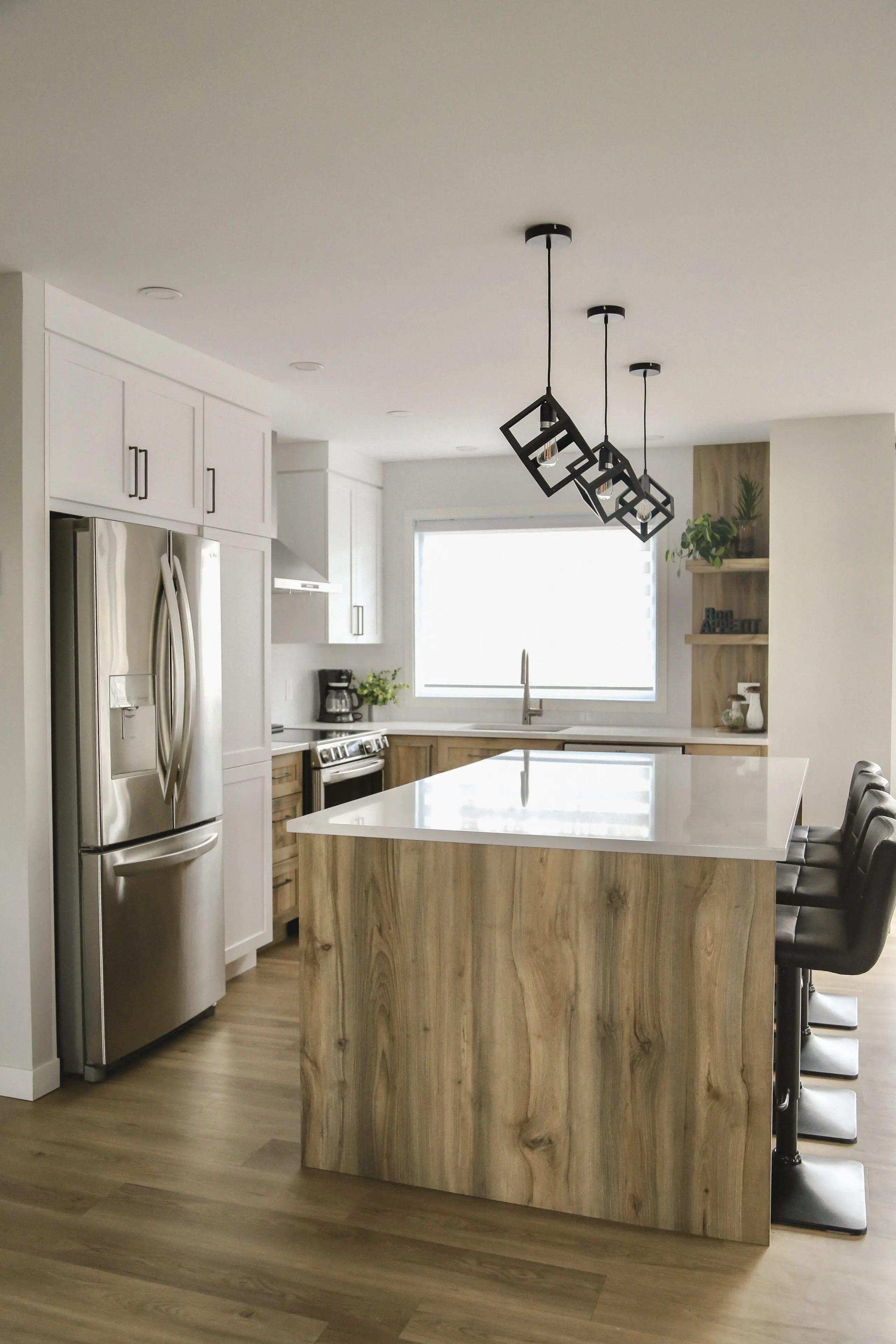 Cuisine moderne avec un grand îlot en bois, des armoires blanches, des appareils en acier inoxydable, des lampes suspendues noires, des tabourets de bar et une grande fenêtre laissant entrer la lumière naturelle.