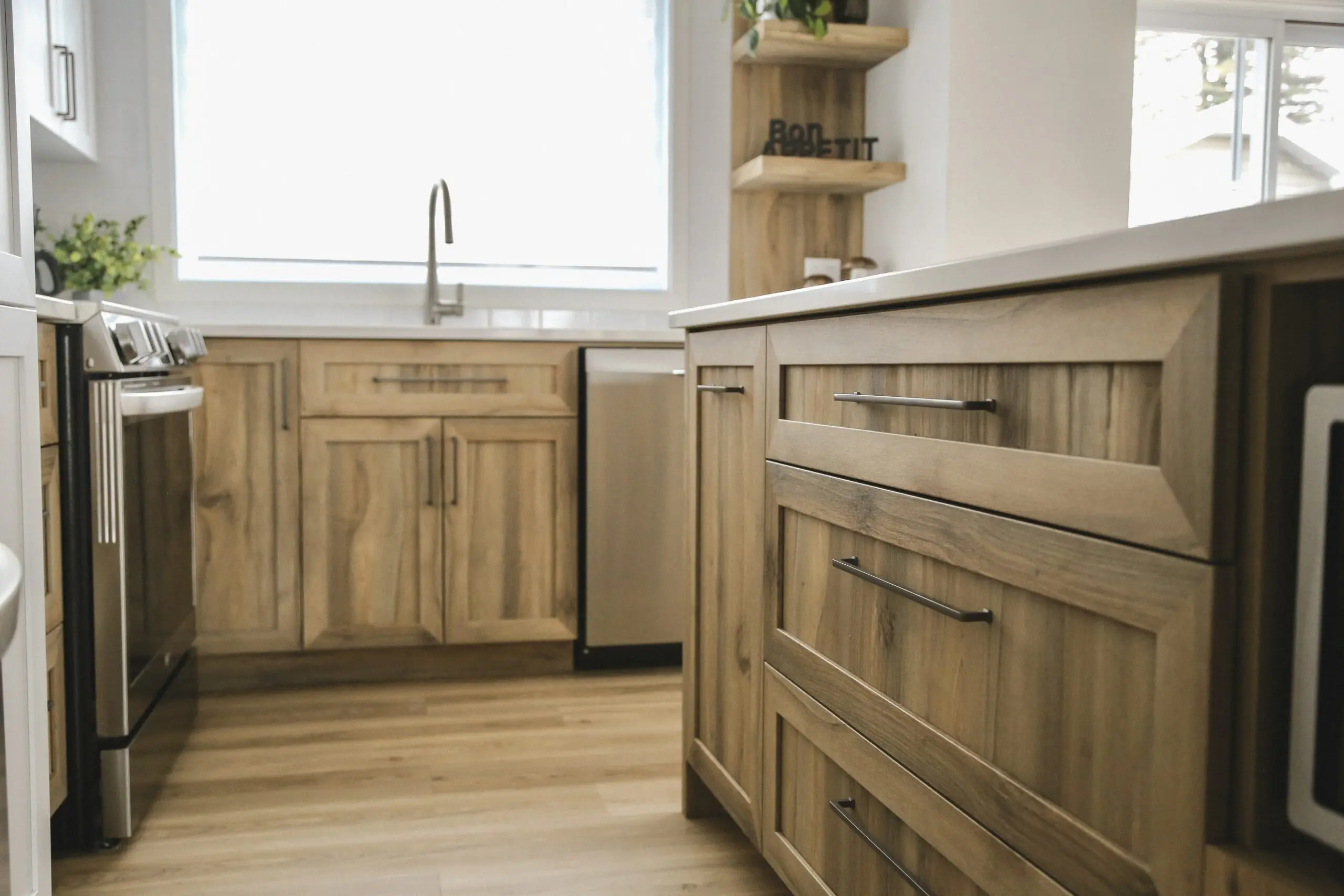 Cuisine moderne avec des armoires et des tiroirs en bois clair, des appareils électroménagers en acier inoxydable, un évier sous une grande fenêtre et des plantes en pot sur le comptoir et l'étagère. La lumière naturelle remplit l'espace.