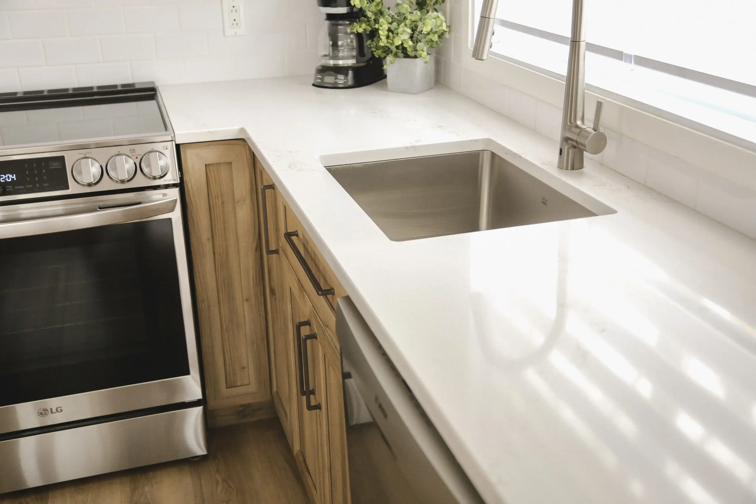 Cuisine moderne avec comptoir blanc, cuisinière en acier inoxydable, four encastré, évier de style maison de campagne, armoires en bois, cafetière et plante en pot près d'une grande fenêtre laissant entrer la lumière naturelle.