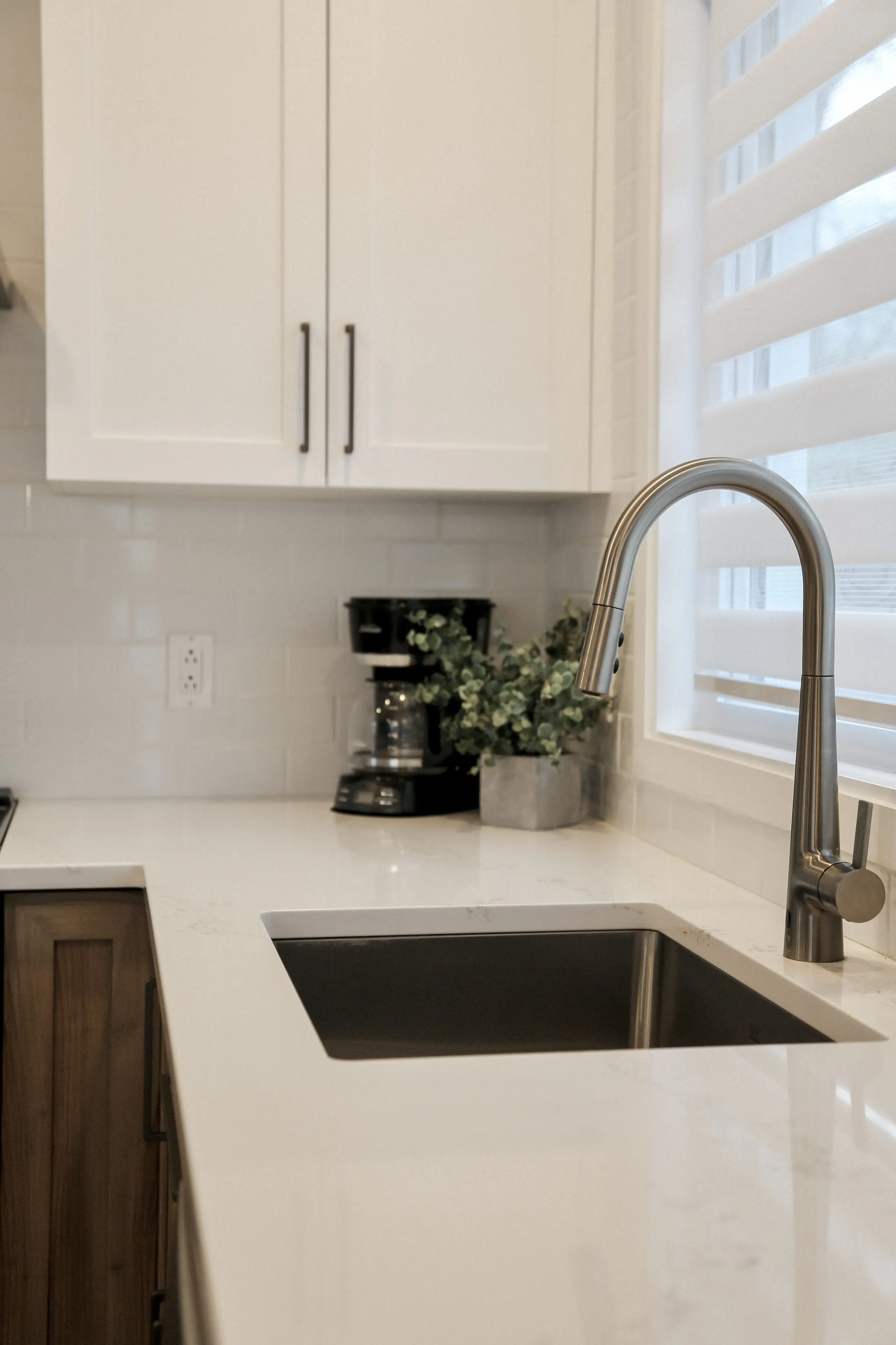 Une cuisine moderne avec des armoires blanches, un évier et un robinet en acier inoxydable, une cafetière et une petite plante en pot sur le plan de travail, près d'une fenêtre aux stores blancs.