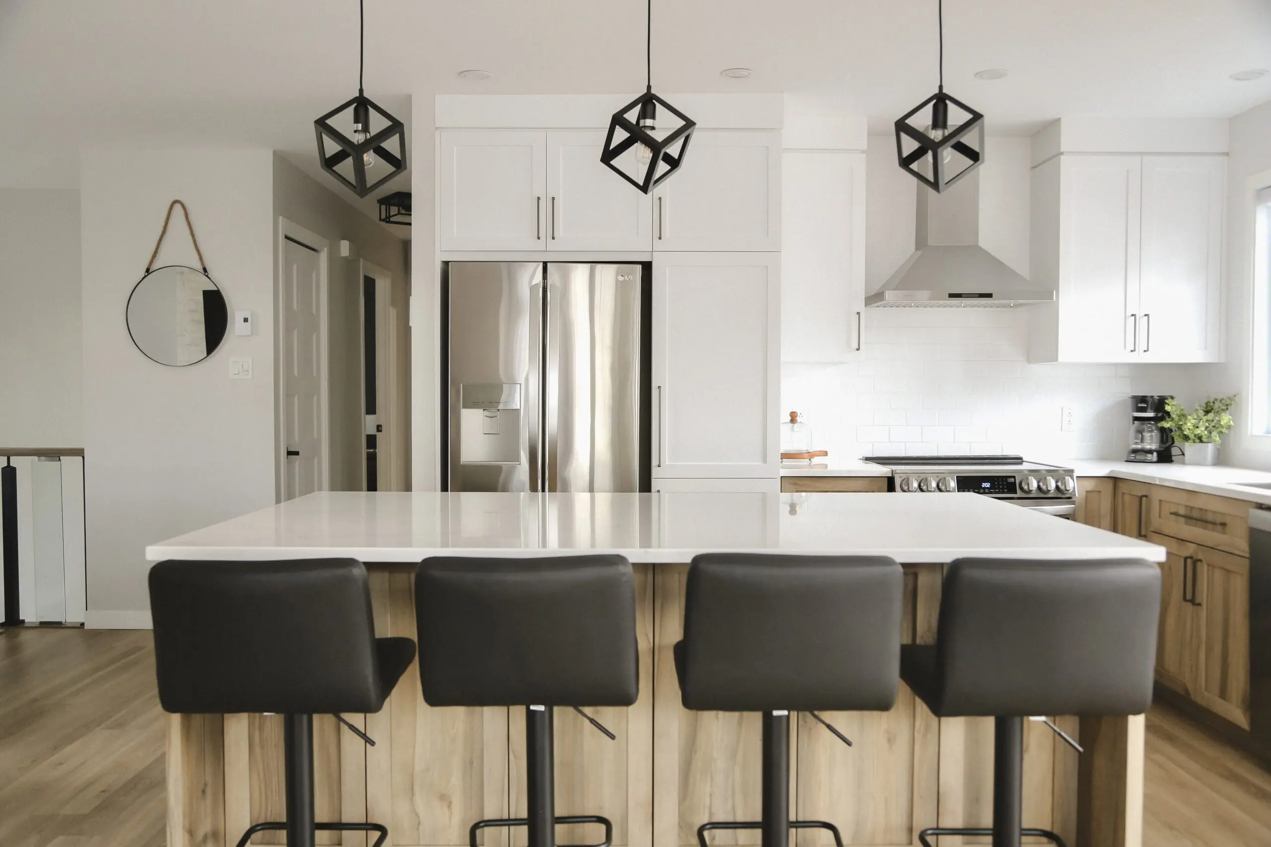 Cuisine moderne avec des armoires blanches, des appareils en acier inoxydable, un grand îlot avec quatre tabourets de bar noirs, des lampes suspendues géométriques et des touches de bois clair. Un miroir rond est accroché au mur à gauche.