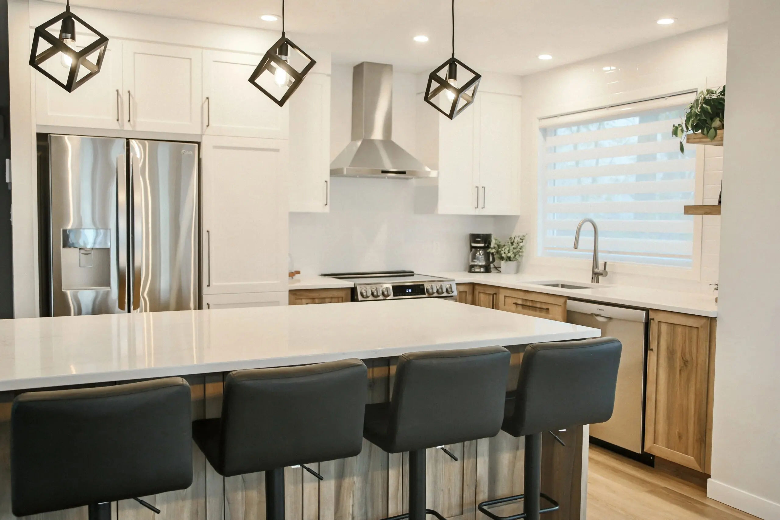 Cuisine moderne avec des armoires blanches, des appareils en acier inoxydable, un îlot central avec quatre tabourets de bar noirs, des lampes suspendues géométriques, une grande fenêtre avec des stores et des accents en bois sur les armoires inférieures.