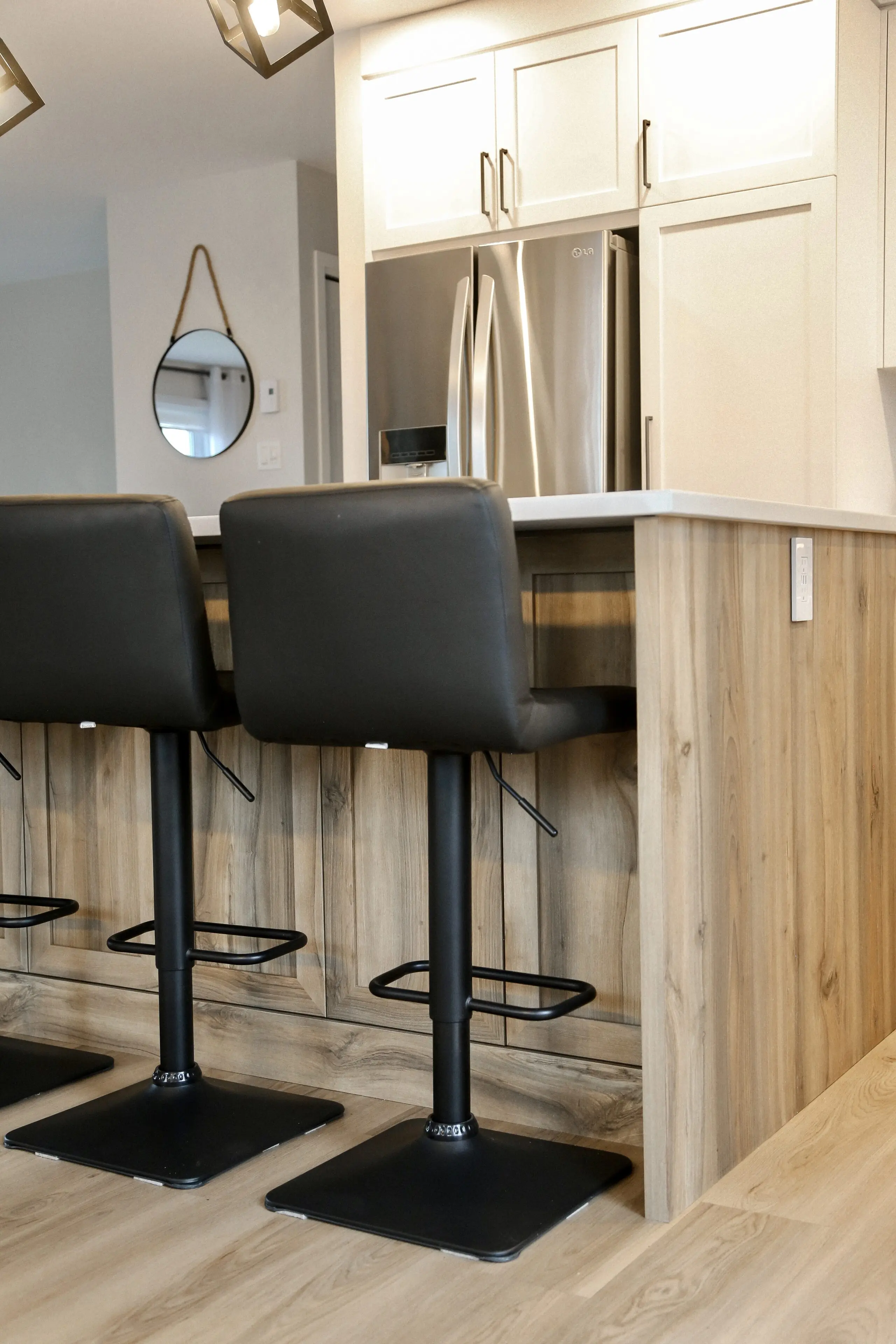 Une cuisine moderne avec un îlot en bois, deux tabourets de bar noirs et un réfrigérateur en acier inoxydable. Un sol en bois clair et un miroir rond sont visibles à l'arrière-plan.
