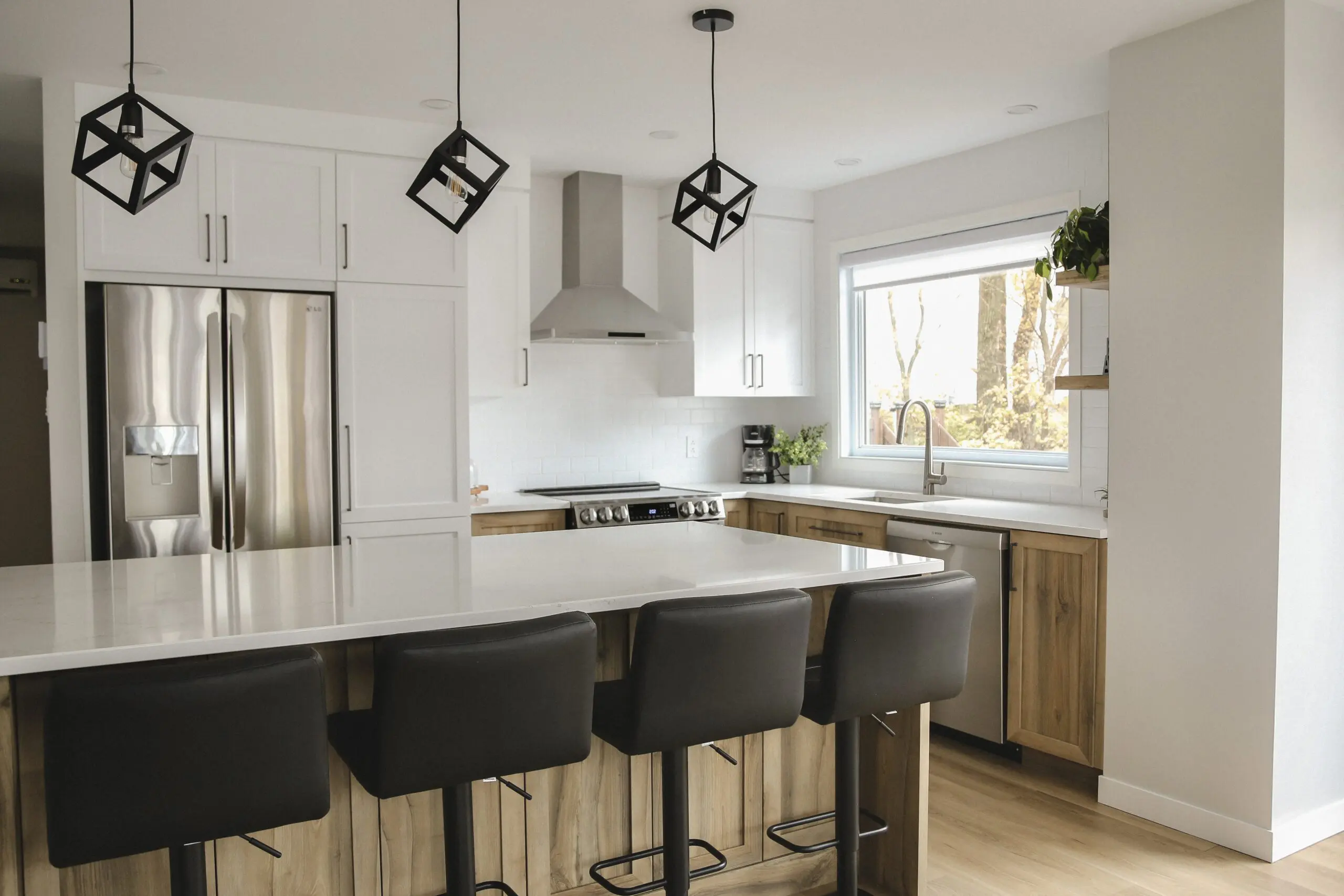 Cuisine moderne avec des armoires blanches, des appareils en acier inoxydable, un grand îlot avec quatre tabourets de bar noirs, des accents en bois, des lampes suspendues, une fenêtre au-dessus de l'évier et des plantes en pot en guise de décor.