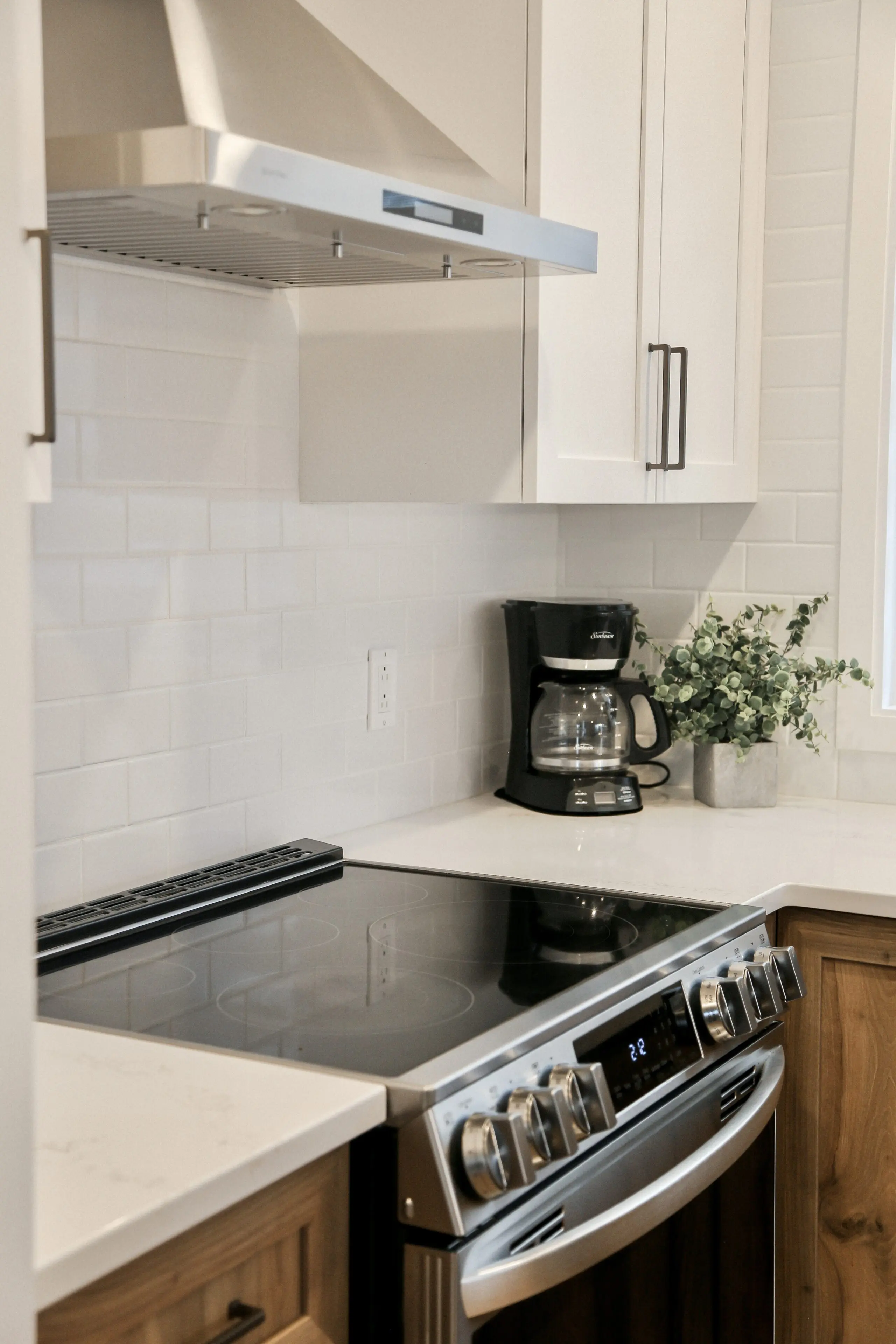 Cuisine moderne avec une cuisinière électrique en acier inoxydable, des armoires blanches, une cafetière, une plante en pot et une hotte en acier inoxydable contre un dosseret en carreaux blancs.