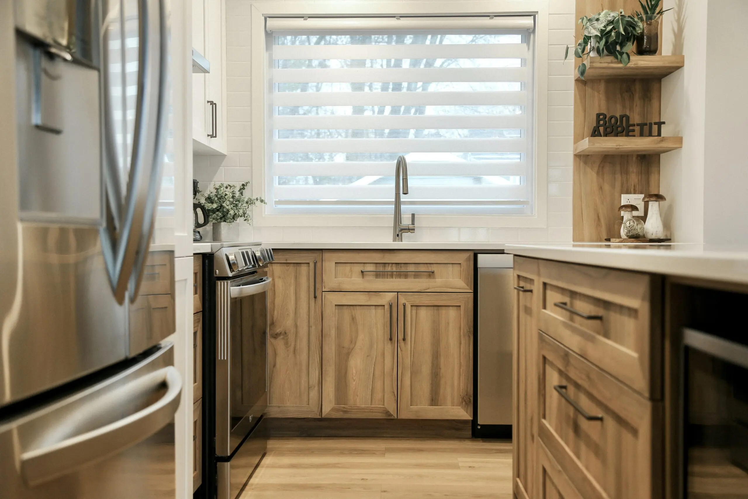 Cuisine moderne avec des armoires en bois clair, des appareils en acier inoxydable, une grande fenêtre avec des stores, un robinet à col de cygne, des comptoirs blancs et des plantes comme décor sur le comptoir et l'étagère.
