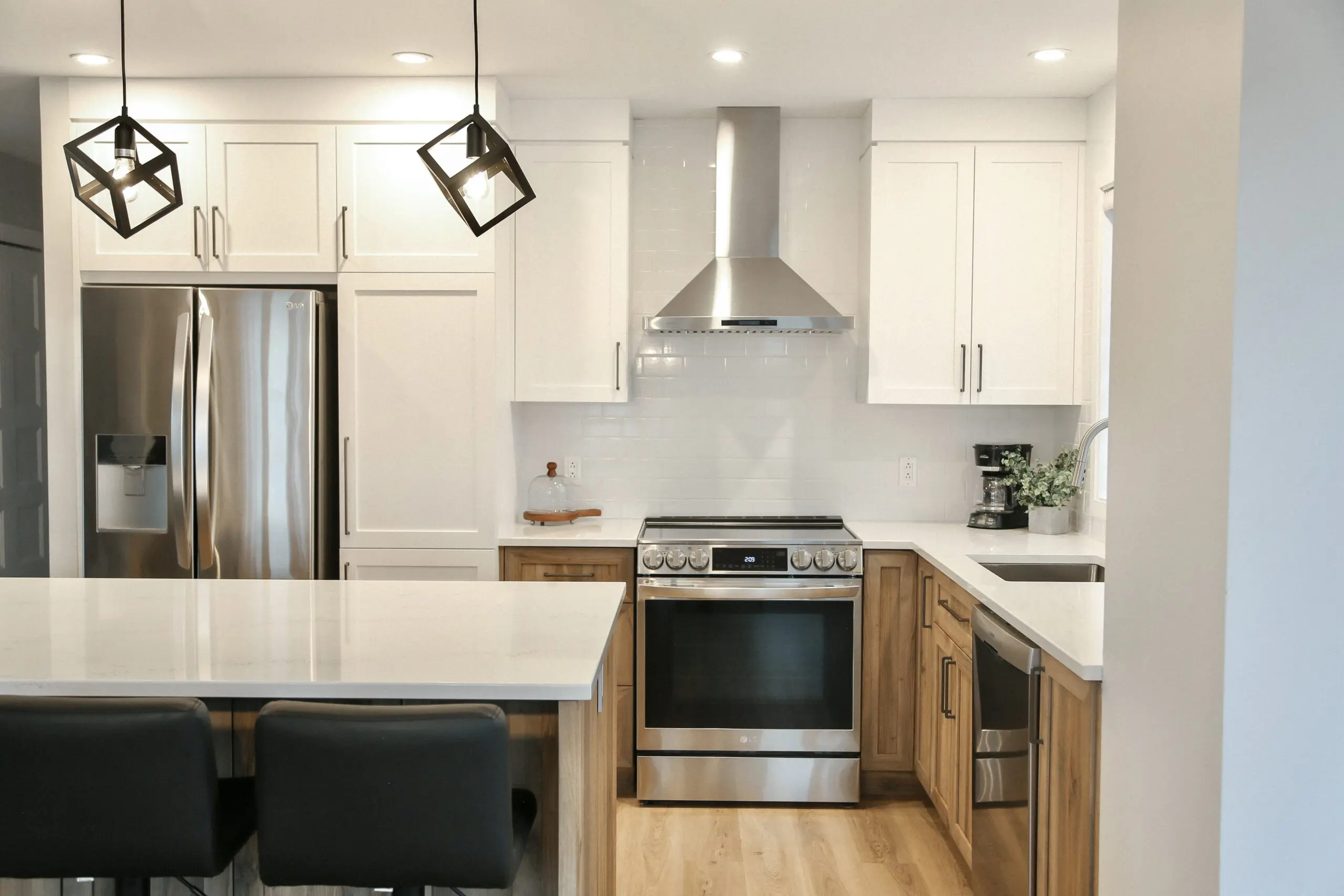 Cuisine moderne avec des armoires blanches, des appareils en acier inoxydable, des accents en bois, un îlot central avec des chaises noires, des lampes suspendues géométriques et une cafetière sur le comptoir à côté de l'évier.