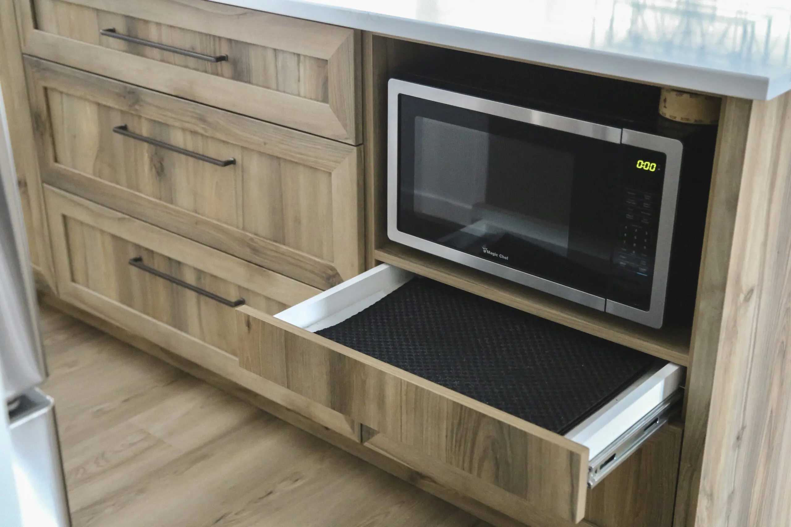 Un meuble de cuisine moderne avec un four à micro-ondes intégré et un tiroir ouvert en dessous, montrant un tapis antidérapant noir à l'intérieur du tiroir. Le meuble est en bois clair avec des poignées en métal.