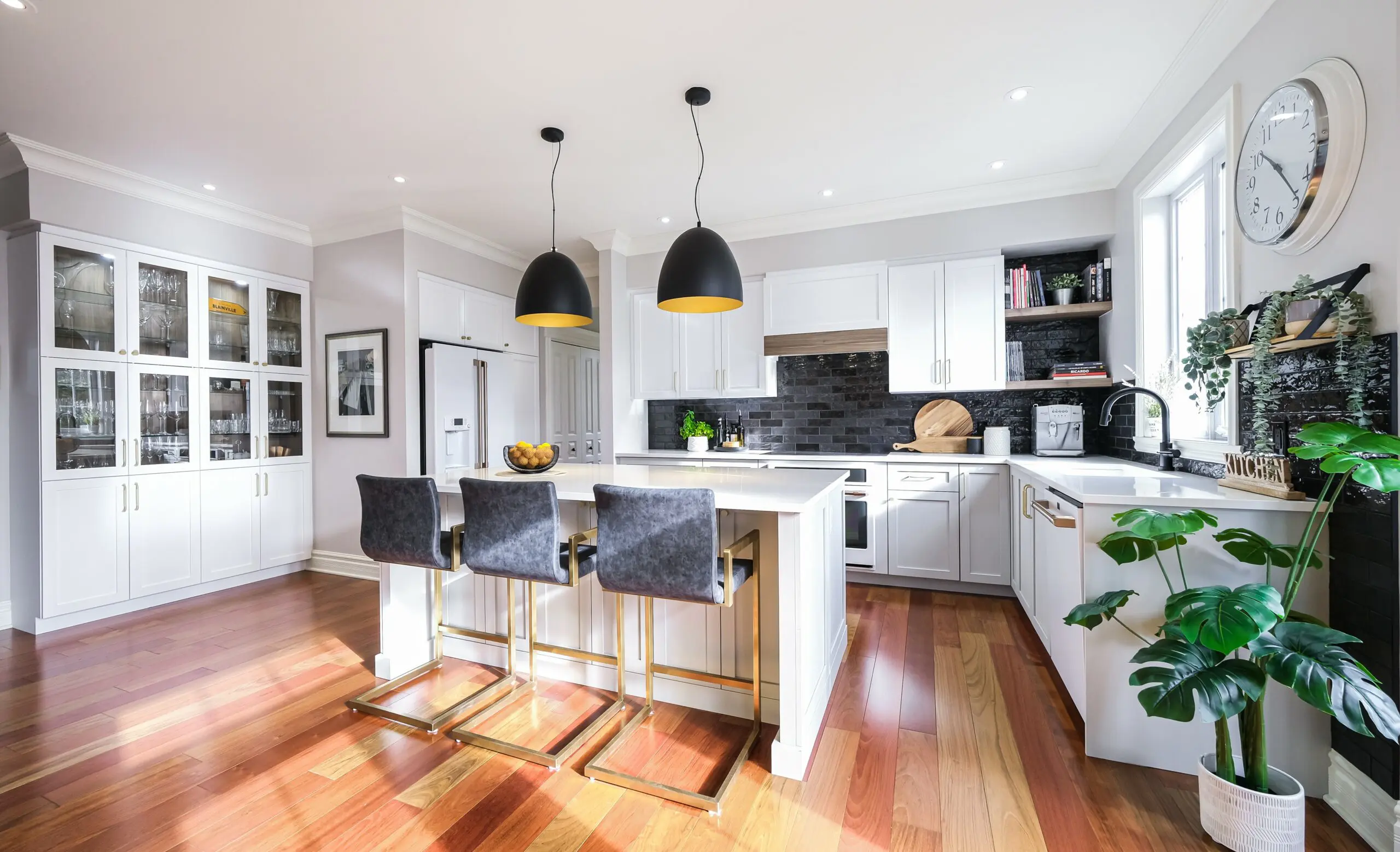 Cuisine moderne et lumineuse avec armoires blanches, dosseret noir, îlot avec trois tabourets de bar noirs, lampes suspendues, sols en bois, plantes en pot et grande fenêtre laissant entrer la lumière naturelle.