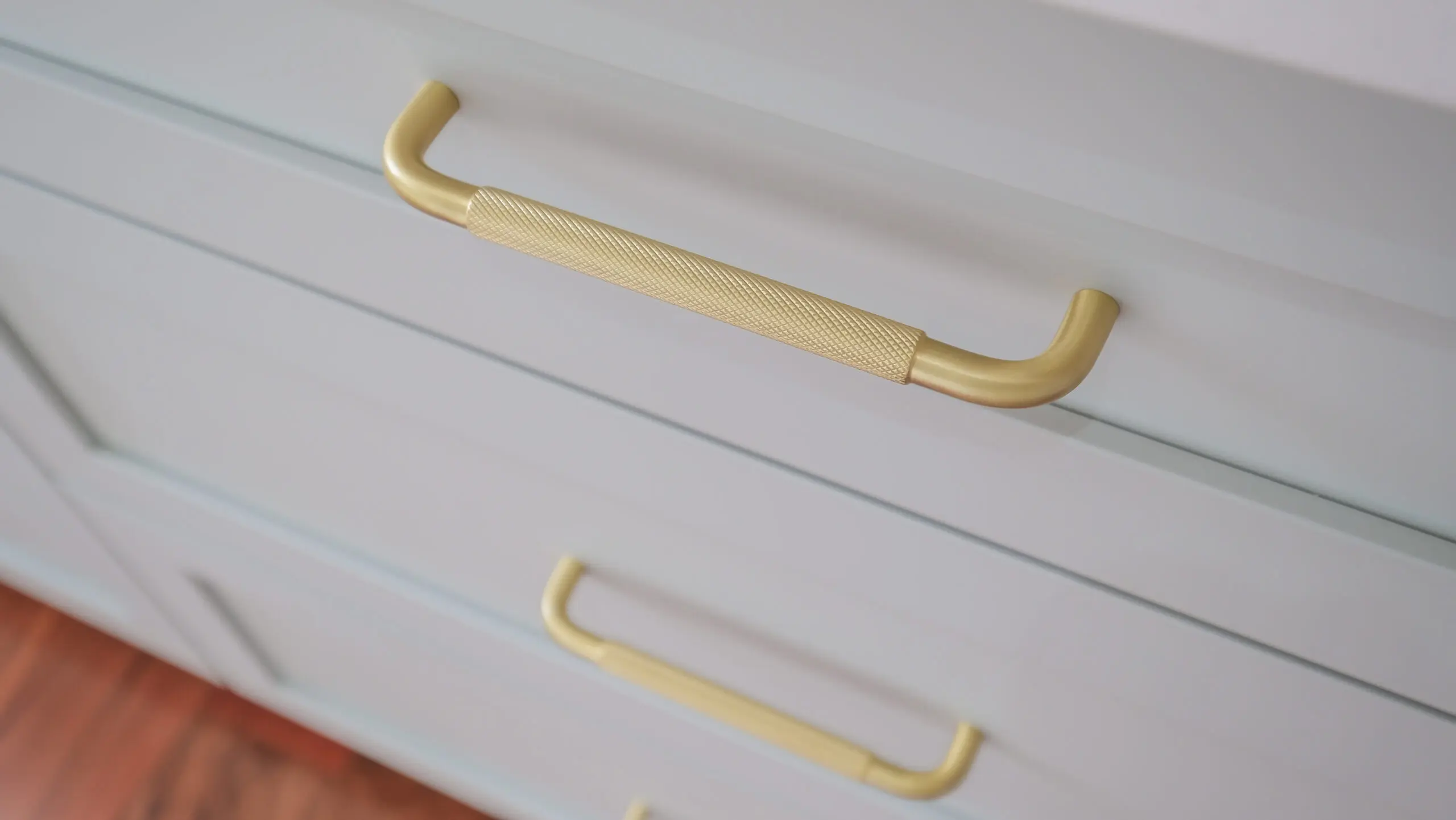 Gros plan sur des tiroirs modernes gris clair avec des poignées en métal doré, au design texturé. Les tiroirs font partie d'une armoire ou d'une commode, et les poignées ont un aspect élégant et contemporain.