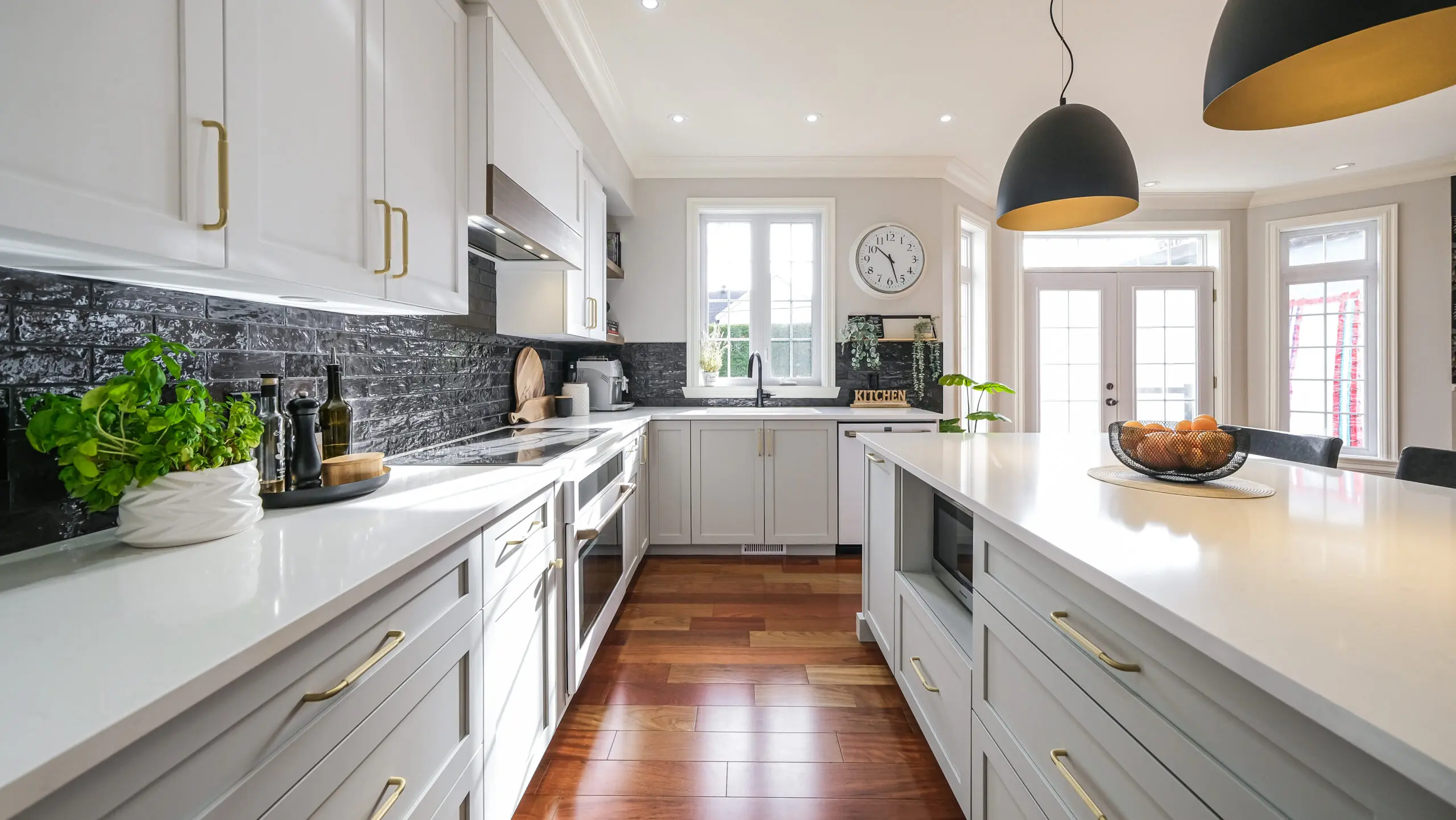 Cuisine moderne avec des armoires blanches, des poignées dorées et un grand îlot. Dosseret en carreaux noirs, lampes suspendues et bol d'oranges sur le comptoir. Les fenêtres et les portes vitrées laissent entrer la lumière du soleil. Le plancher en bois ajoute de la chaleur.