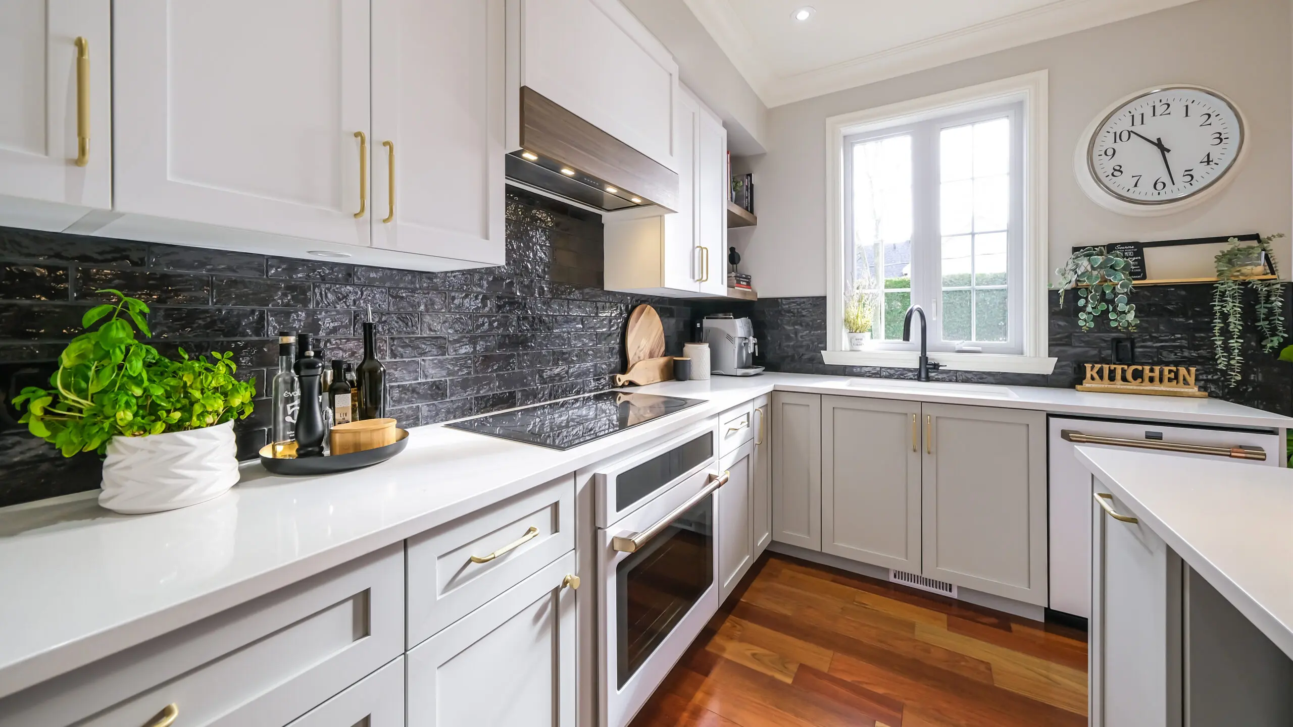 Cuisine moderne avec placards blancs, dosseret en carrelage noir, four encastré et table de cuisson électrique. Une grande fenêtre laisse entrer la lumière naturelle. Les comptoirs accueillent des herbes, des bouteilles et de la décoration de cuisine. Une horloge murale est suspendue au-dessus de l'évier.
