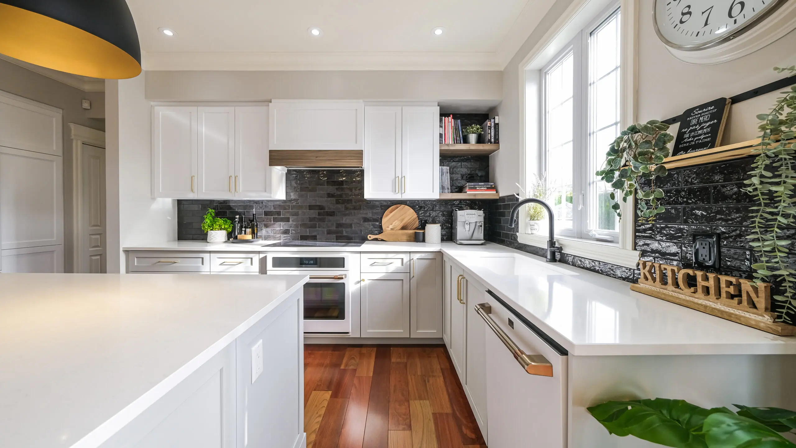 Cuisine moderne avec des armoires blanches, un dosseret en carreaux noirs, des appareils en acier inoxydable, un plancher en bois, de grandes fenêtres, des plantes décoratives et des ustensiles de cuisine sur les comptoirs et les étagères. Espace lumineux, propre et organisé.