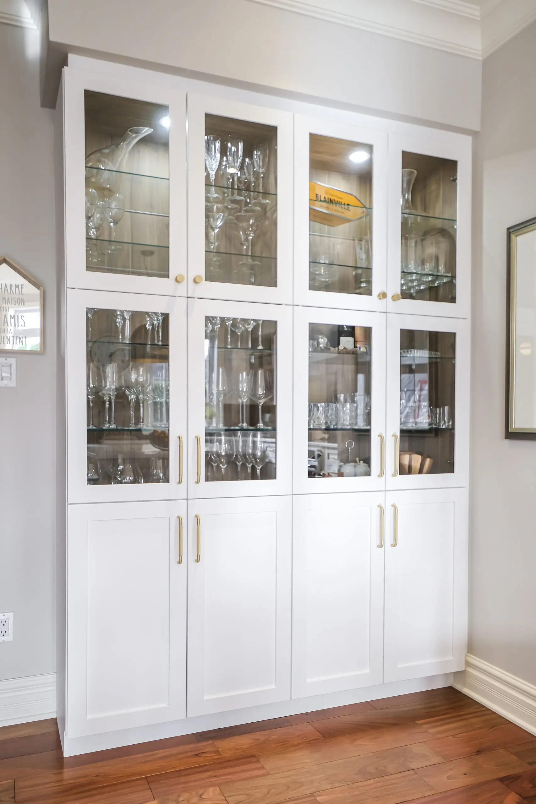 Une armoire encastrée blanche avec des portes en verre présente divers articles en verre, notamment des verres à vin, des flûtes à champagne et des gobelets, sur des étagères en bois. L'armoire est posée sur un sol en bois contre un mur gris clair.