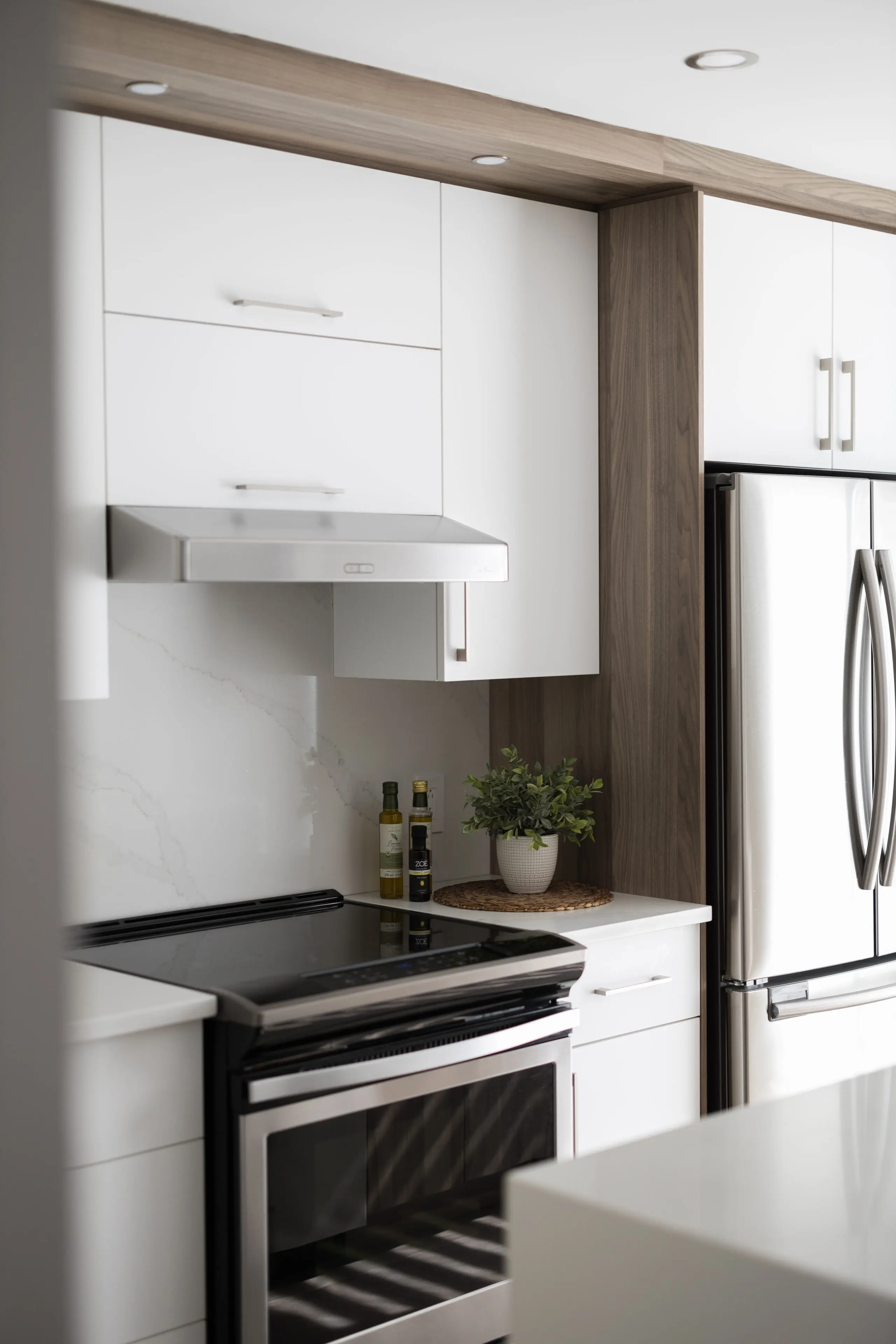 Cuisine moderne avec des armoires blanches, un four et une hotte en acier inoxydable, un réfrigérateur argenté à double porte, une plante en pot et des bouteilles d'huile sur un comptoir blanc avec un dosseret en marbre.