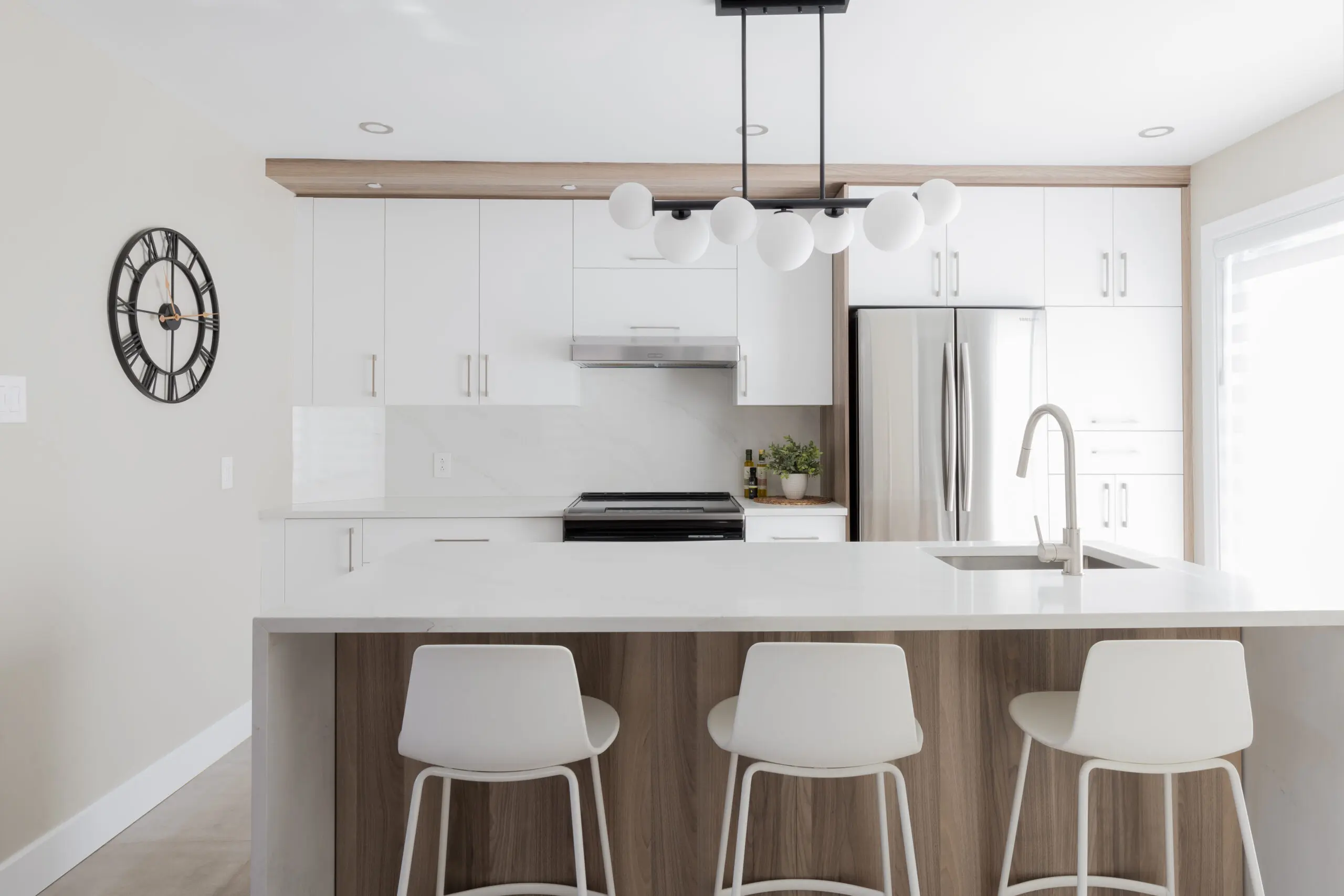 Cuisine moderne avec des armoires blanches, un réfrigérateur en acier inoxydable, un grand îlot avec trois chaises blanches, une horloge minimaliste en noir et blanc sur le mur et un luminaire contemporain au-dessus de l'îlot.