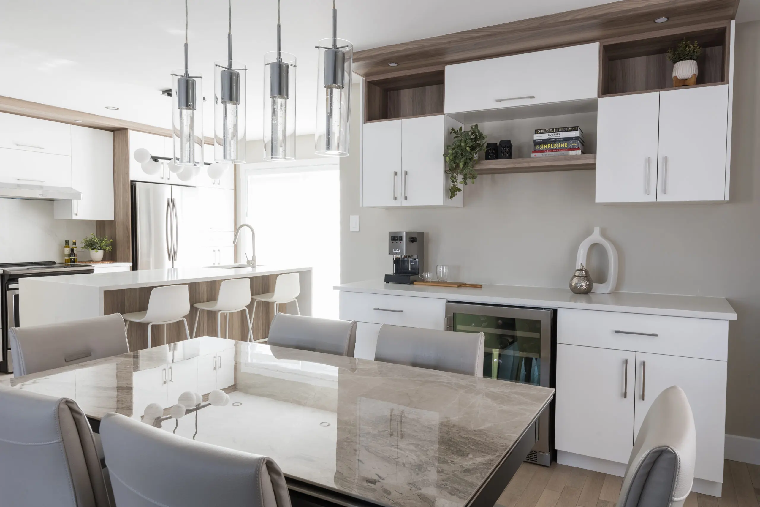Cuisine et salle à manger modernes avec table en marbre, chaises grises, armoires blanches, lampes suspendues et îlot de cuisine avec tabourets de bar. Des étagères accueillent des livres et des plantes ; un refroidisseur de vin et une cafetière sont posés sur le comptoir.