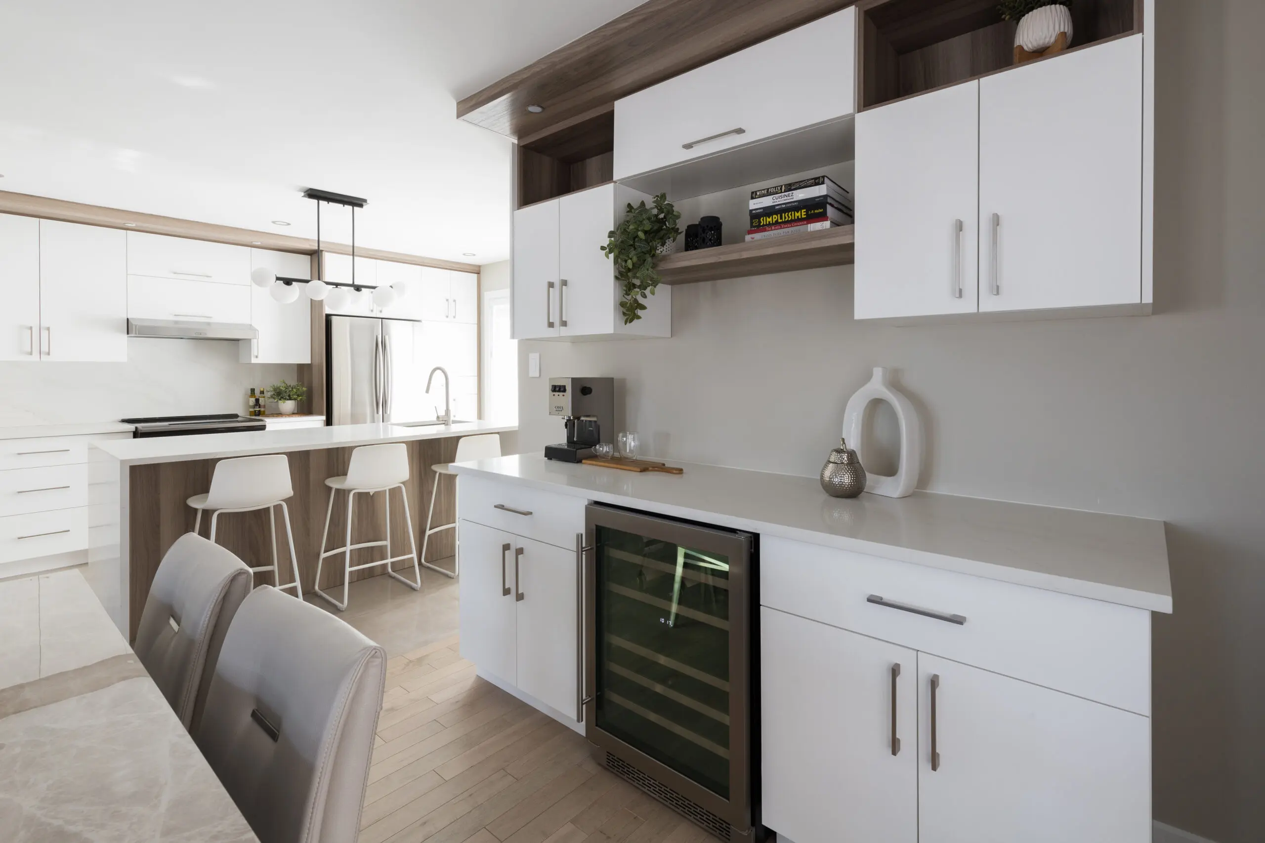 Cuisine moderne avec des armoires blanches, un réfrigérateur à vin, des étagères ouvertes, une cafetière et des tabourets de bar sur un îlot. Des plantes décoratives et des livres ajoutent des accents à cet espace clair et épuré.