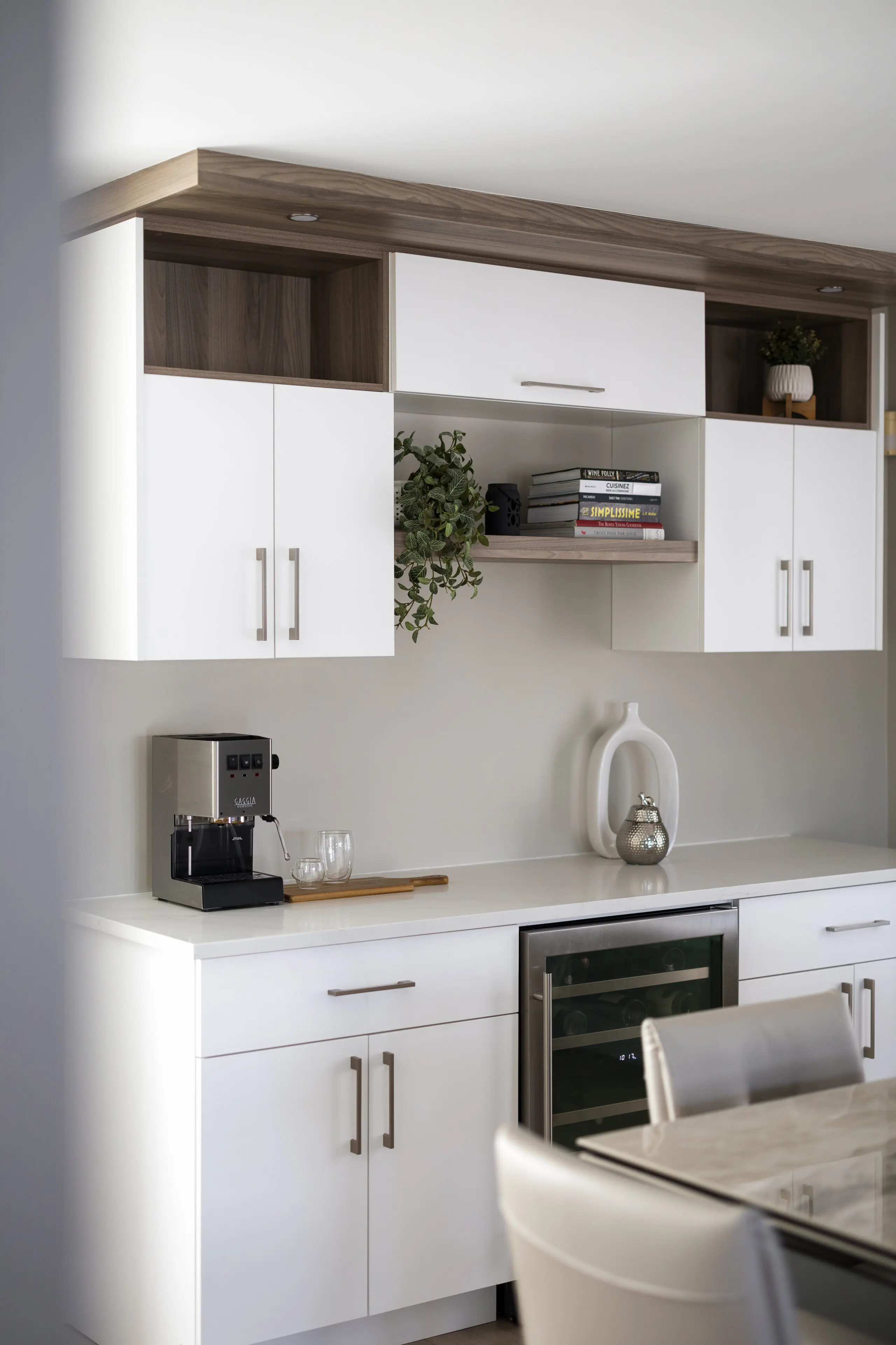 Cuisine moderne avec des armoires blanches, un comptoir sur lequel se trouvent une machine à café, deux verres, un plateau en bois, un vase décoratif et quelques plantes, avec des livres et des objets de décoration sur des étagères ouvertes au-dessus.