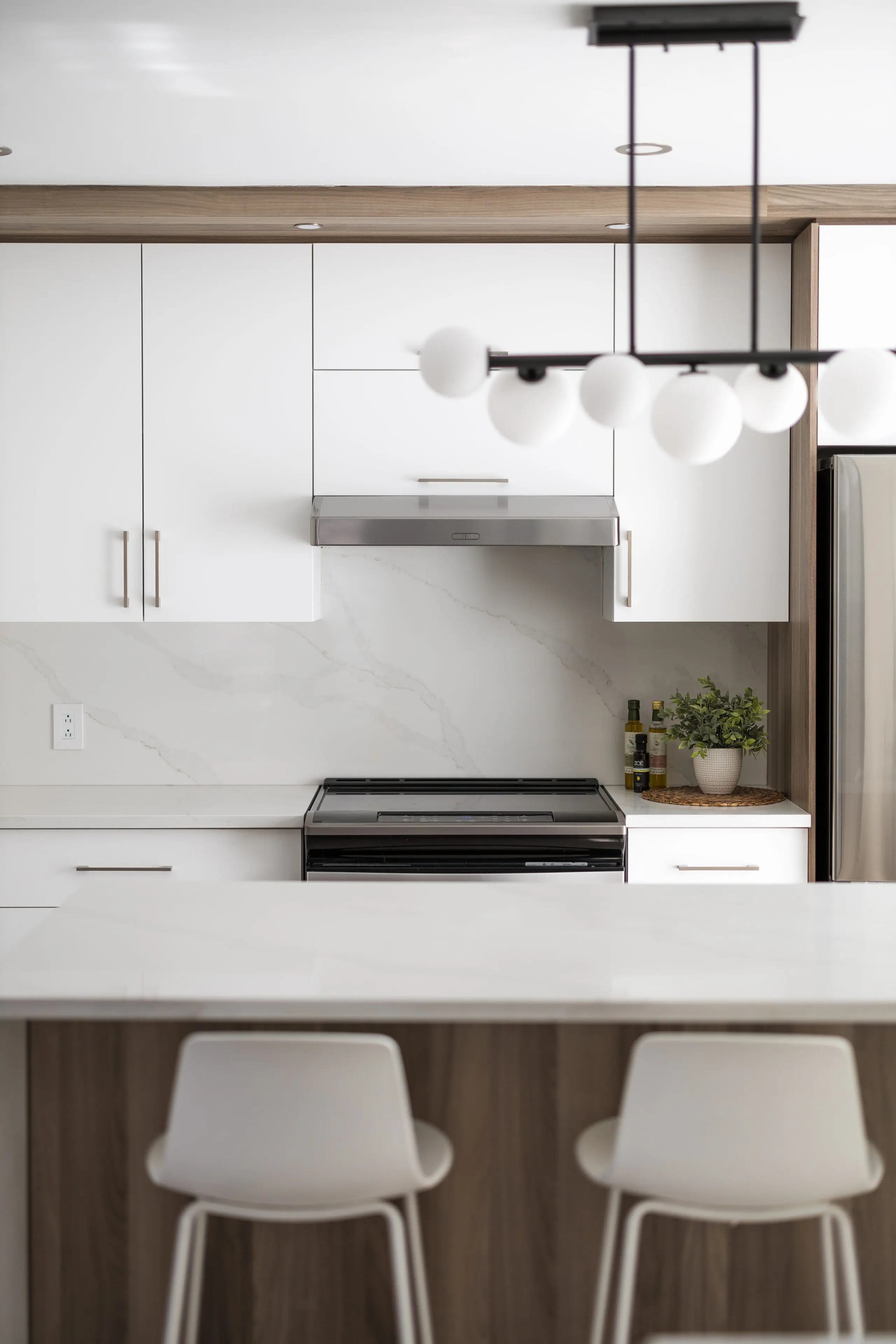 Cuisine moderne avec des armoires blanches, un four et une hotte en acier inoxydable, un dosseret en marbre, deux tabourets de bar blancs sur un comptoir clair, une plante et un luminaire contemporain noir avec des ampoules rondes.