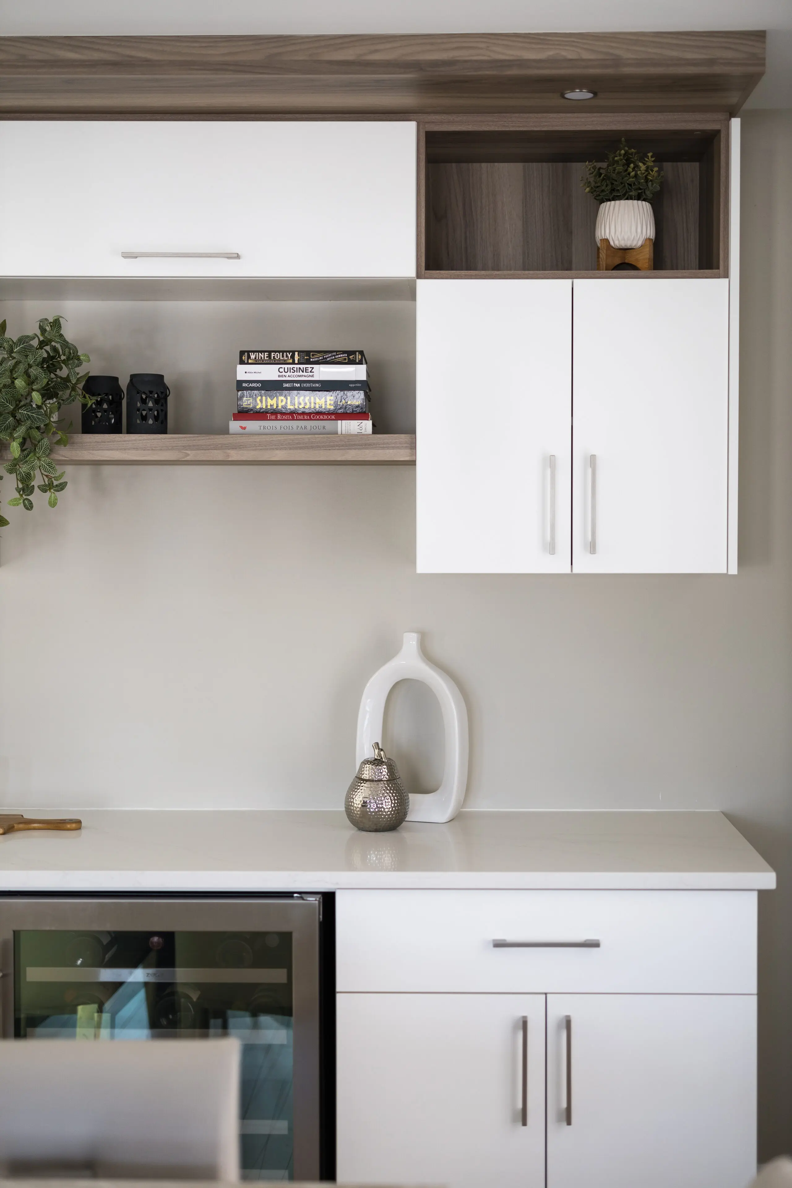Cuisine moderne avec des armoires blanches, une étagère en bois contenant des livres et des plantes, un petit réfrigérateur à vin et des objets décoratifs sur le comptoir, notamment un vase blanc et un ornement métallique en forme de poire.
