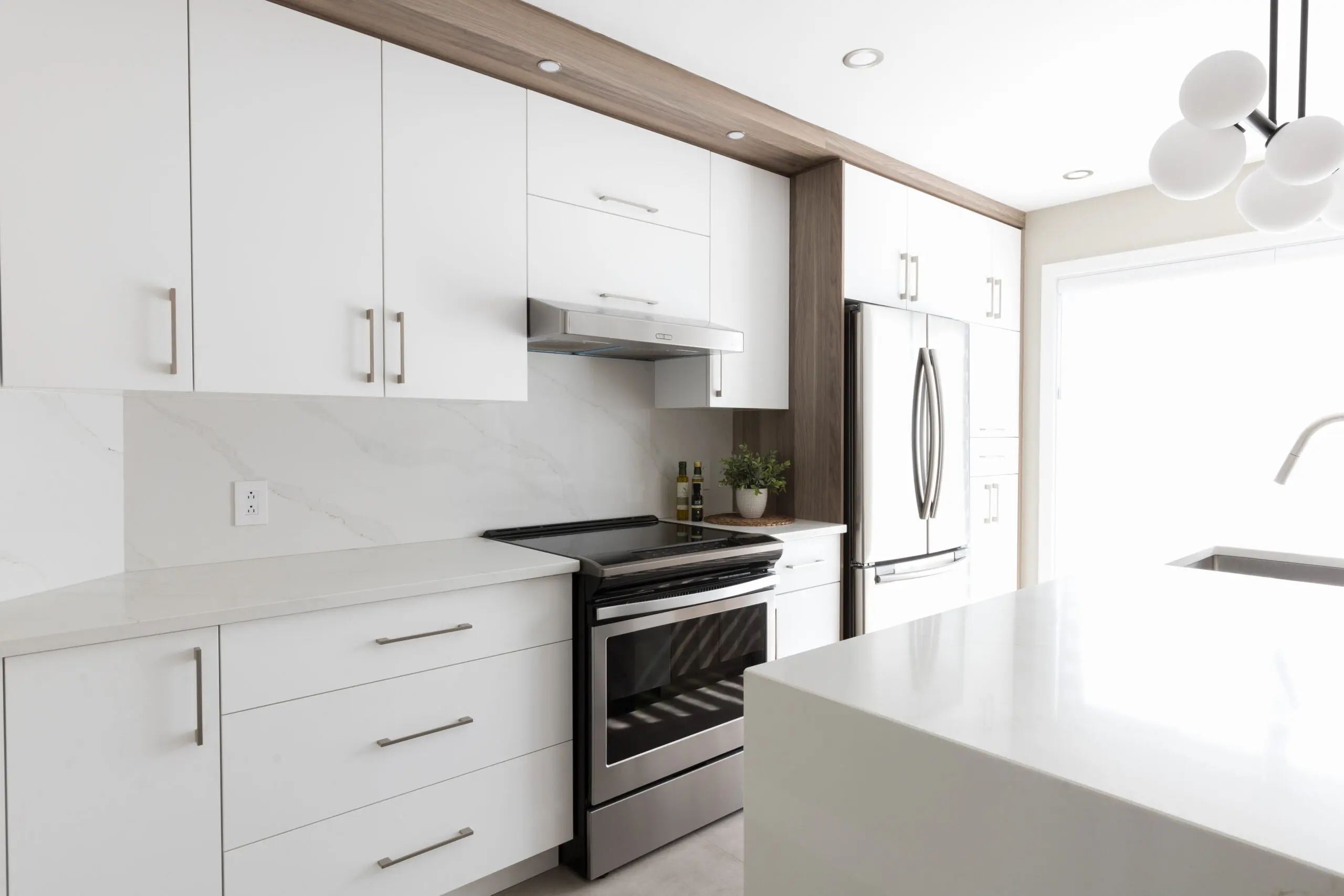 Cuisine moderne avec des armoires blanches, des appareils en acier inoxydable, un dosseret en marbre et un îlot. Une cuisinière avec une hotte est au centre, et un réfrigérateur se trouve à droite, près d'une grande fenêtre.