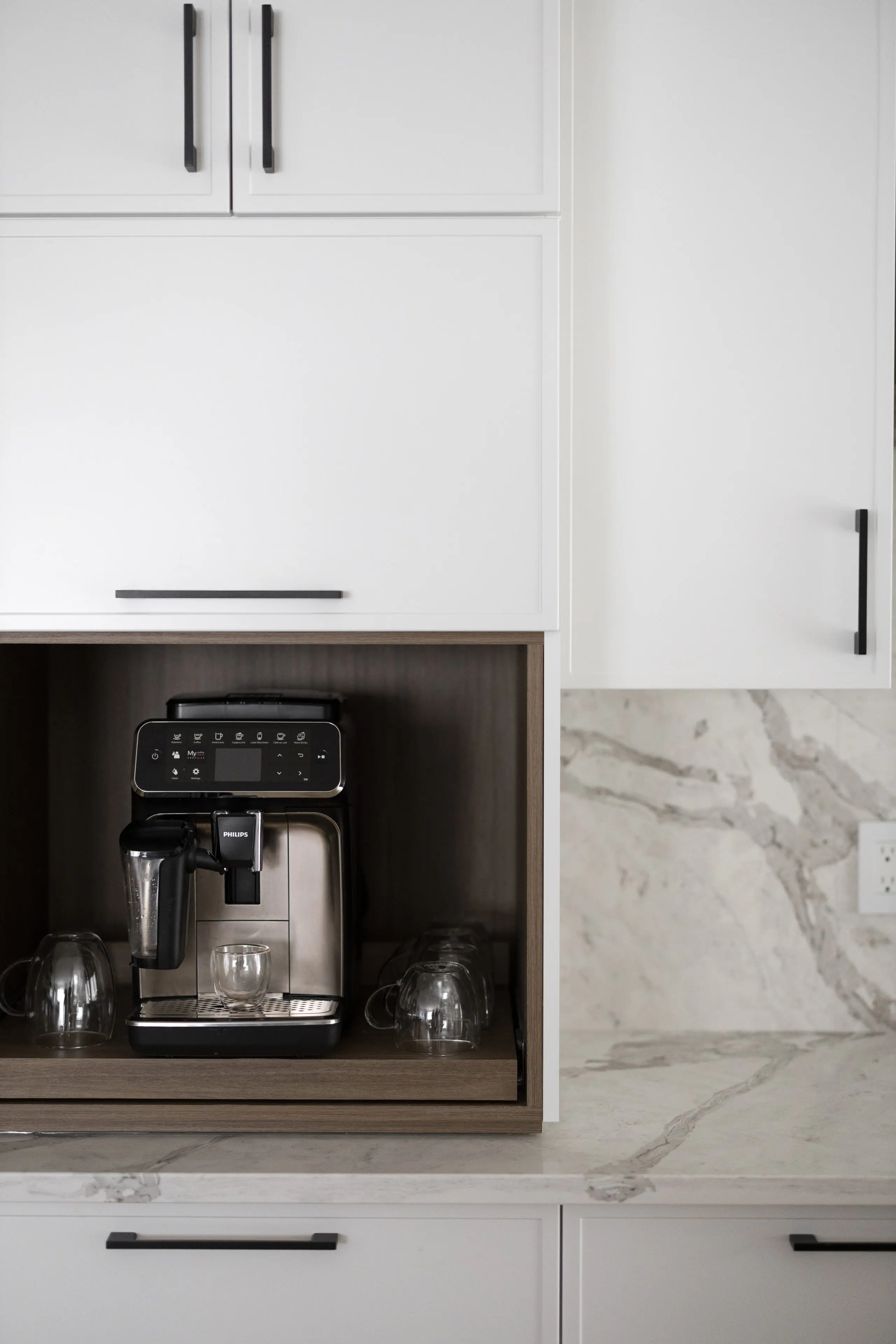 Une machine à café moderne avec des tasses en verre est placée dans un coin encastré dans une cuisine blanche avec des comptoirs en marbre et des poignées d'armoire foncées.