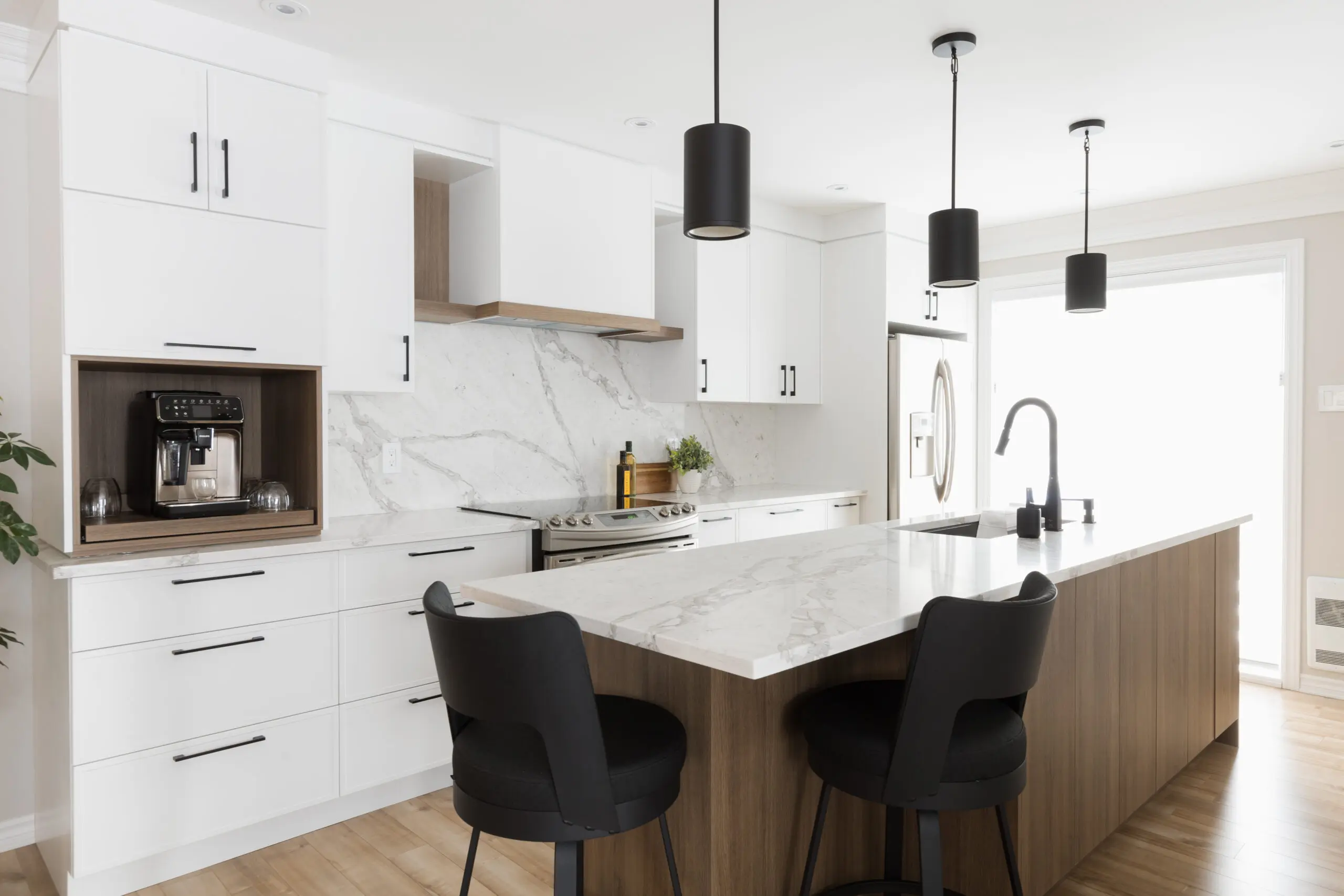 Cuisine moderne avec des armoires blanches, des comptoirs et un dosseret en marbre, une machine à café encastrée, des lampes suspendues noires et deux chaises noires sur un îlot avec un évier et un robinet. Espace lumineux avec des touches de bois.