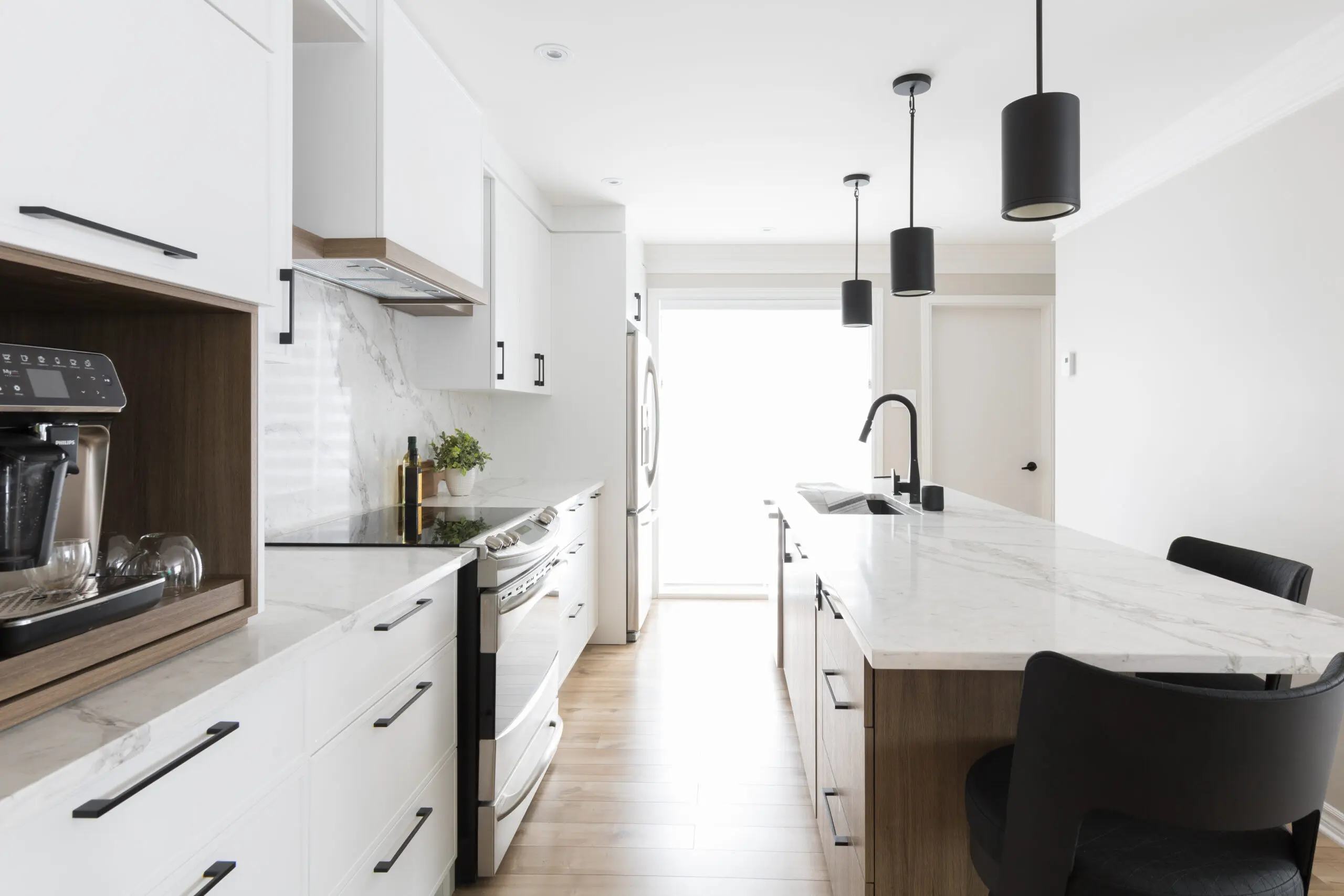 Cuisine moderne avec armoires blanches, comptoirs en marbre, appareils électroménagers en acier inoxydable, cafetière intégrée, lampes suspendues noires, grand îlot avec évier et chaises noires sur un sol en bois clair.