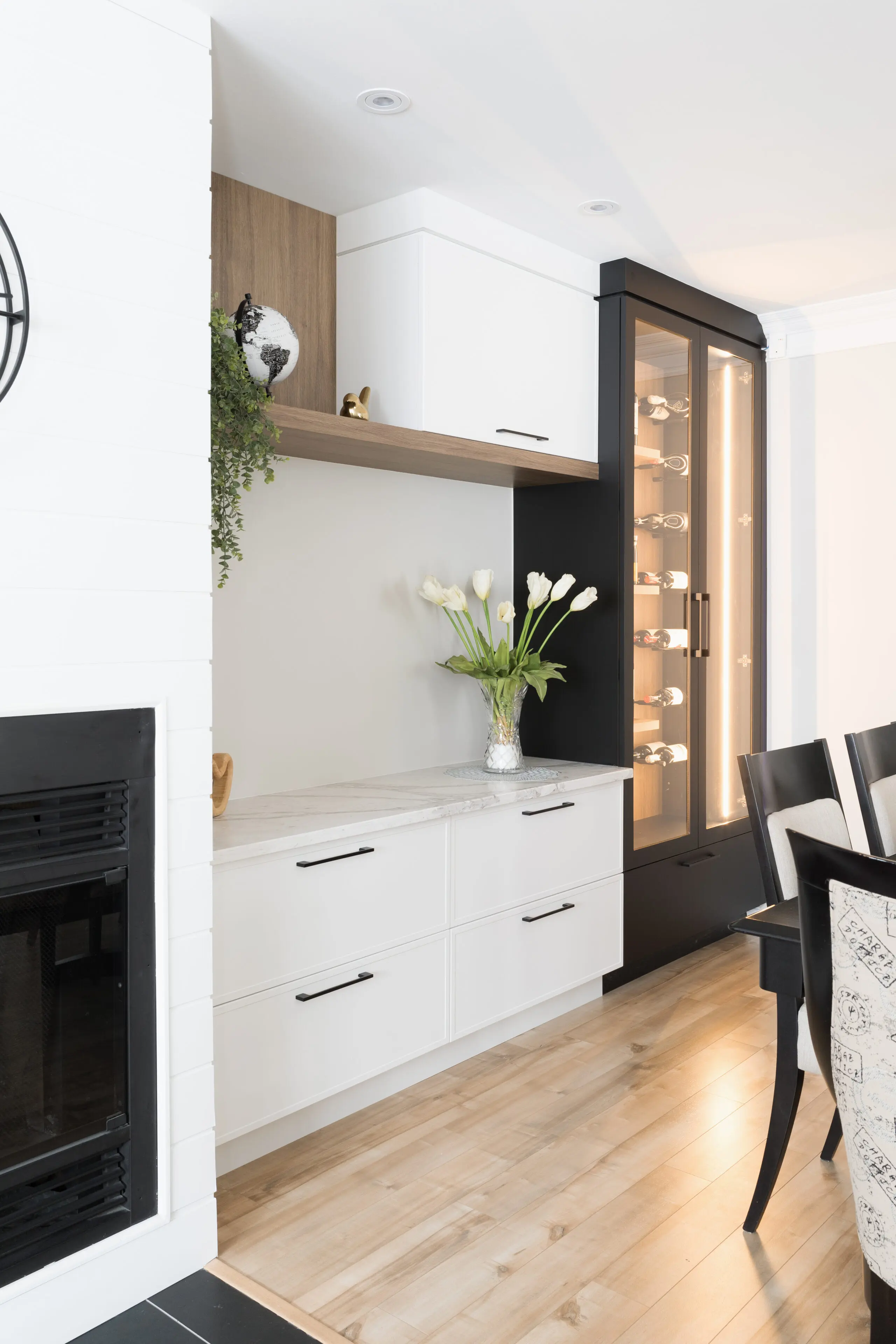 Une salle à manger moderne avec des planchers en bois clair, des armoires blanches, un comptoir en marbre, un vase de tulipes blanches, une vitrine avec des lumières et des chaises de salle à manger noires. Une cheminée est visible sur la gauche.
