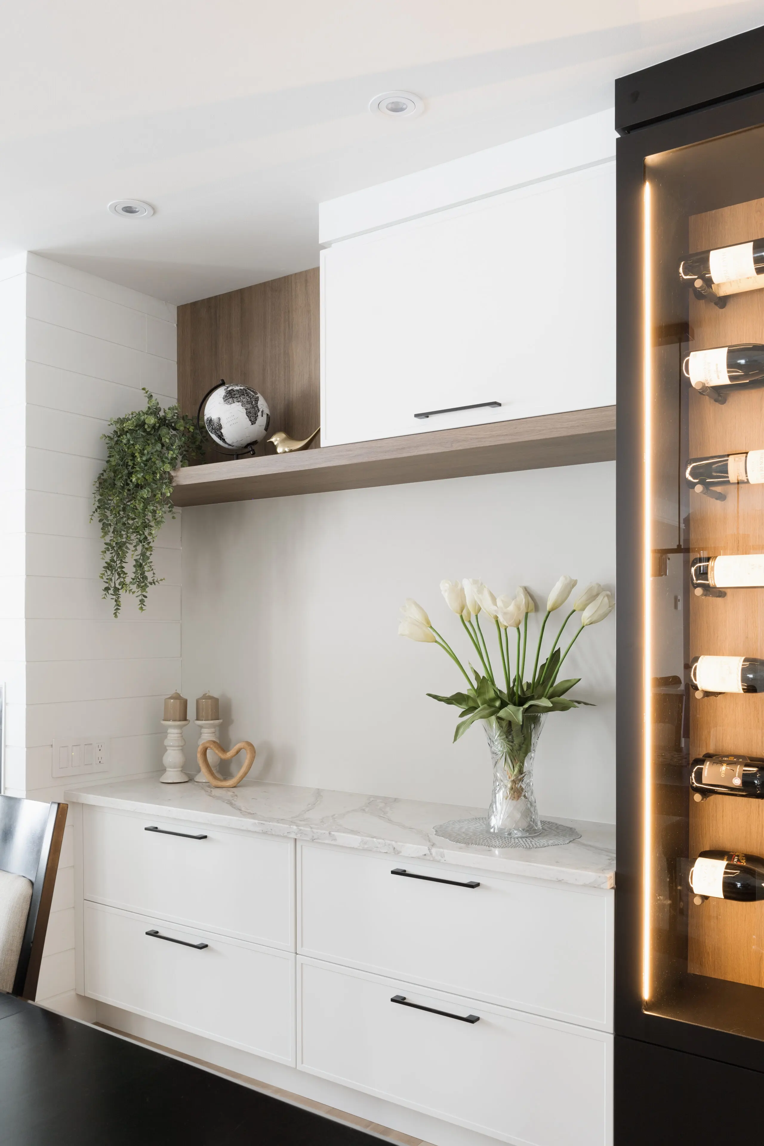 Coin de cuisine moderne avec des armoires blanches, un comptoir en marbre, un vase en verre avec des tulipes blanches, une étagère contenant un globe, des bougies, une plante grimpante et un réfrigérateur à vin éclairé présentant des bouteilles sur la droite.