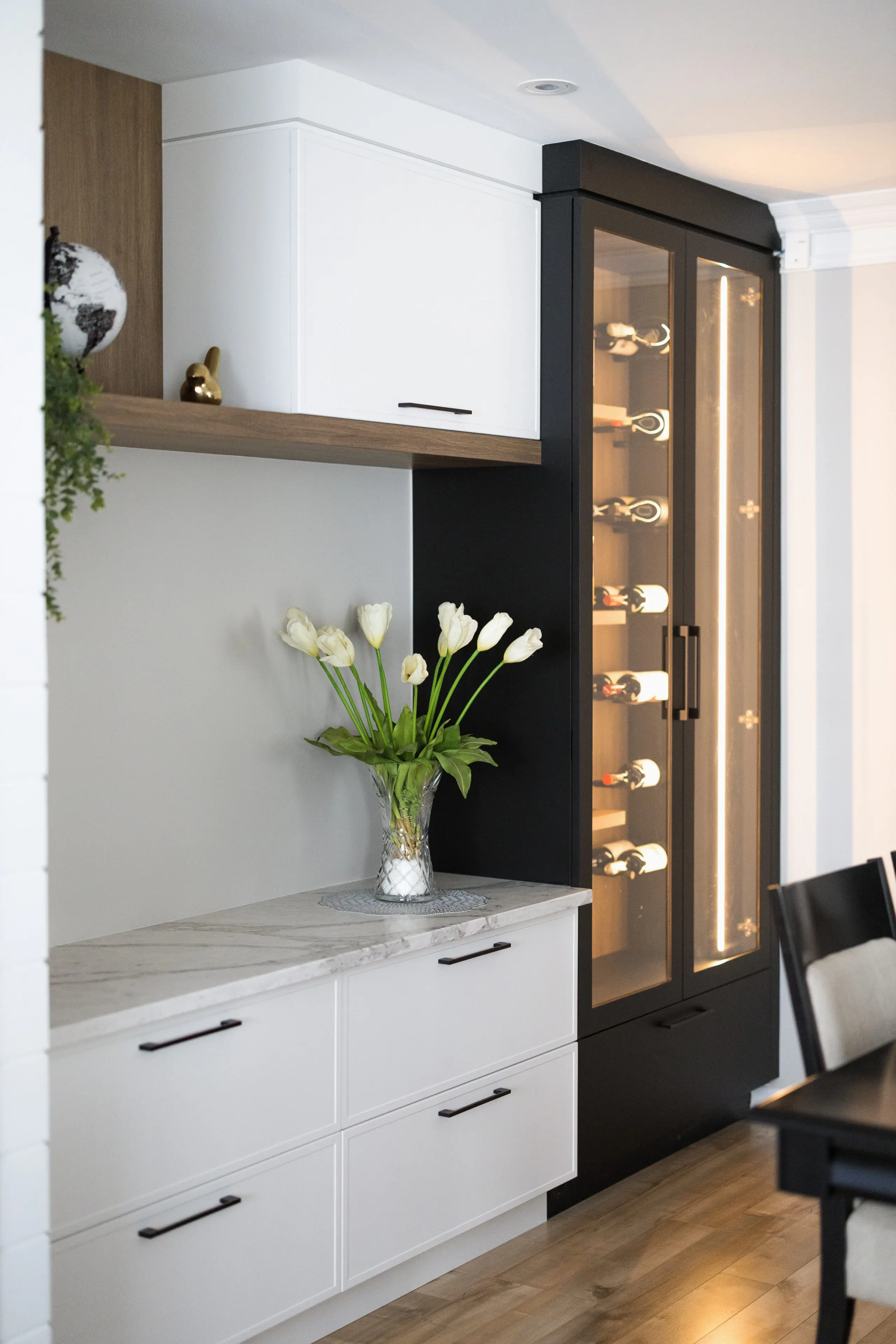 Un coin cuisine moderne avec des armoires blanches, un comptoir en marbre, un vase de tulipes blanches et une haute armoire à vin en verre encadrée de noir et remplie de bouteilles de vin. Le sol en bois et l'éclairage doux créent une atmosphère chaleureuse.
