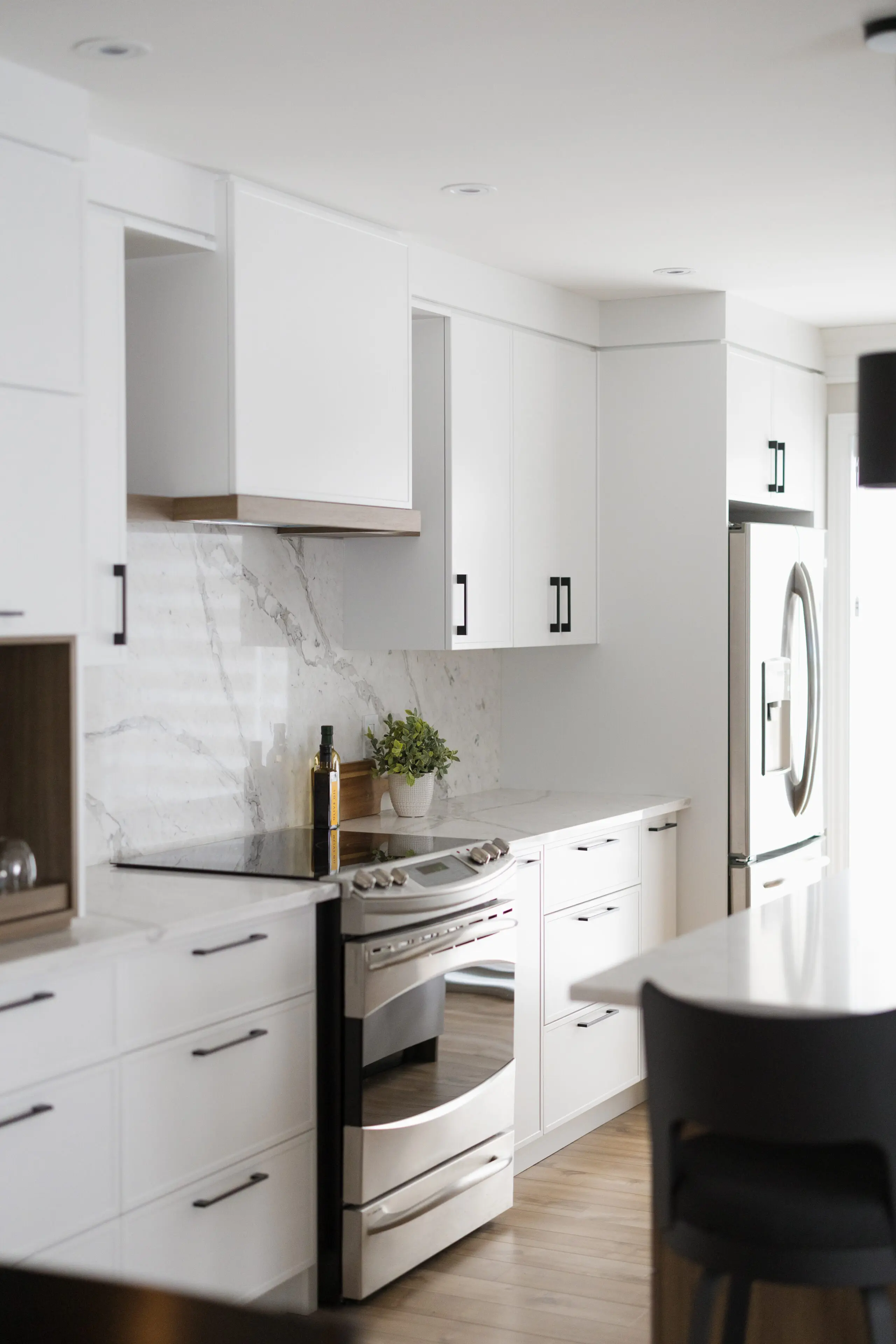 Cuisine moderne avec des armoires blanches, des appareils en acier inoxydable, un dosseret en marbre, une plante en pot, une bouteille d'huile sur le comptoir et un sol en bois. L'espace est baigné de lumière naturelle.