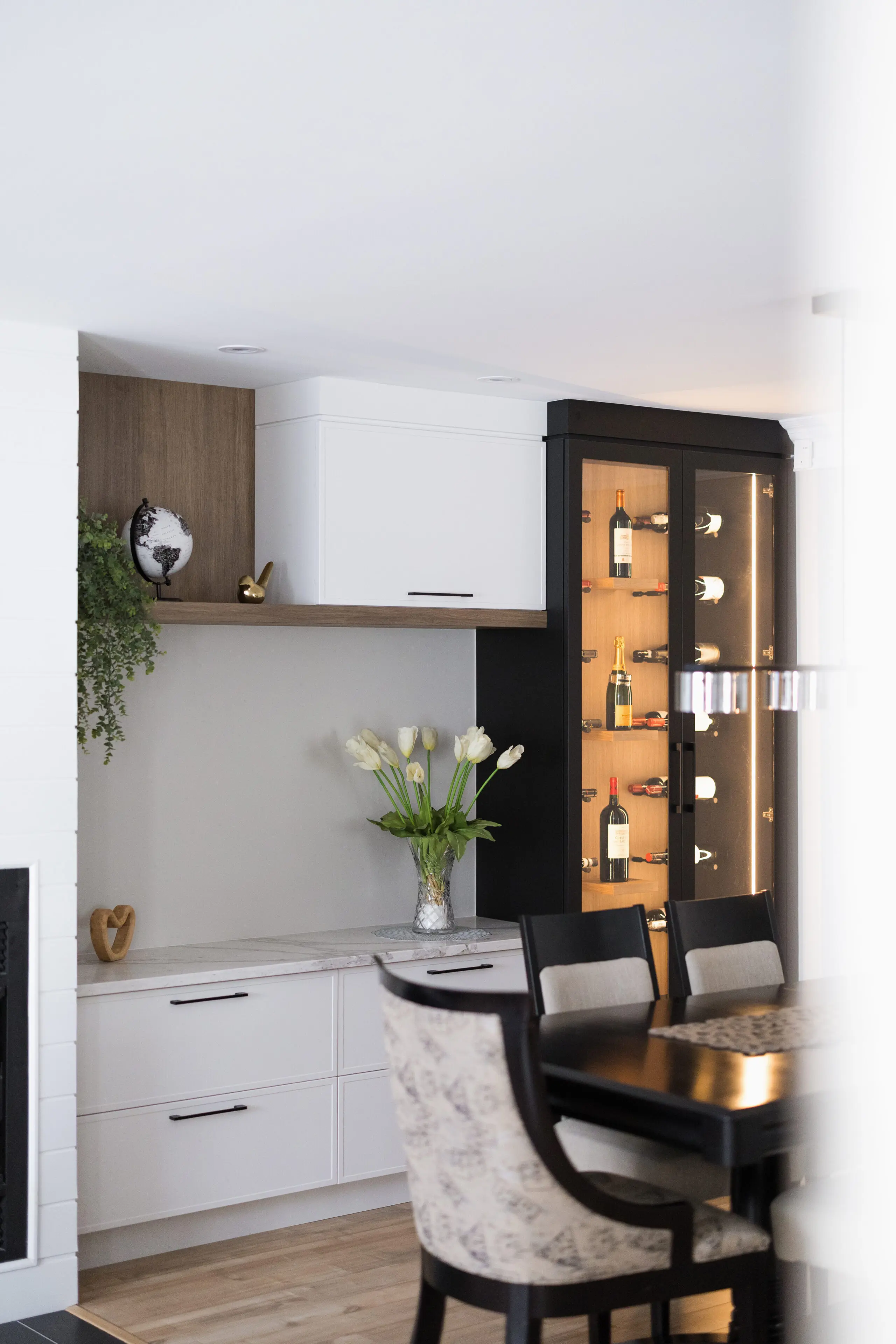 Cuisine et salle à manger modernes avec des armoires blanches, un comptoir en marbre, un vase de tulipes blanches, une plante décorative et une haute armoire vitrée exposant des bouteilles et des verres. Quatre chaises rembourrées entourent une table foncée.