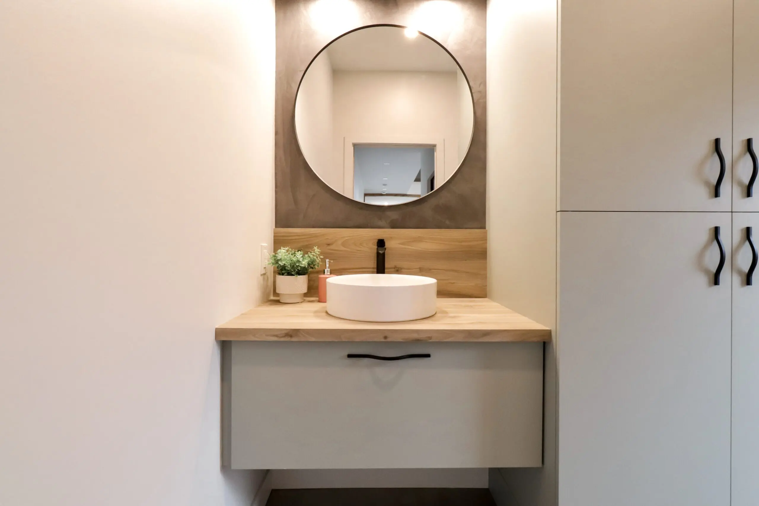 Meuble de salle de bain moderne avec miroir rond, comptoir en bois, évier blanc, robinet noir et plante en pot. Une lumière éclaire le miroir et une armoire avec des poignées noires se trouve à droite.