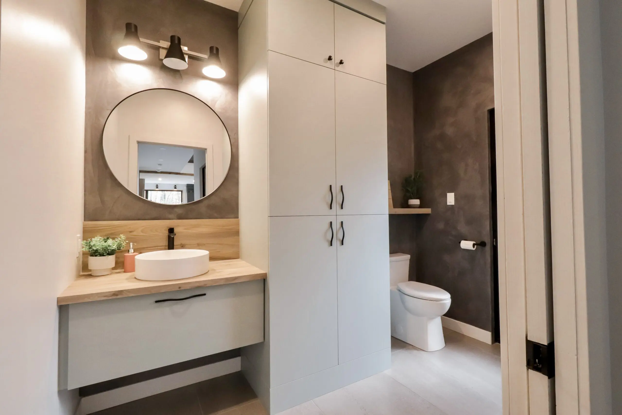 Salle de bains moderne avec un miroir rond au-dessus d'un lavabo sur un plan de travail en bois, des armoires de rangement hautes, des toilettes blanches et des murs gris. Deux appliques murales se trouvent au-dessus du miroir et une petite plante décore le coin lavabo.