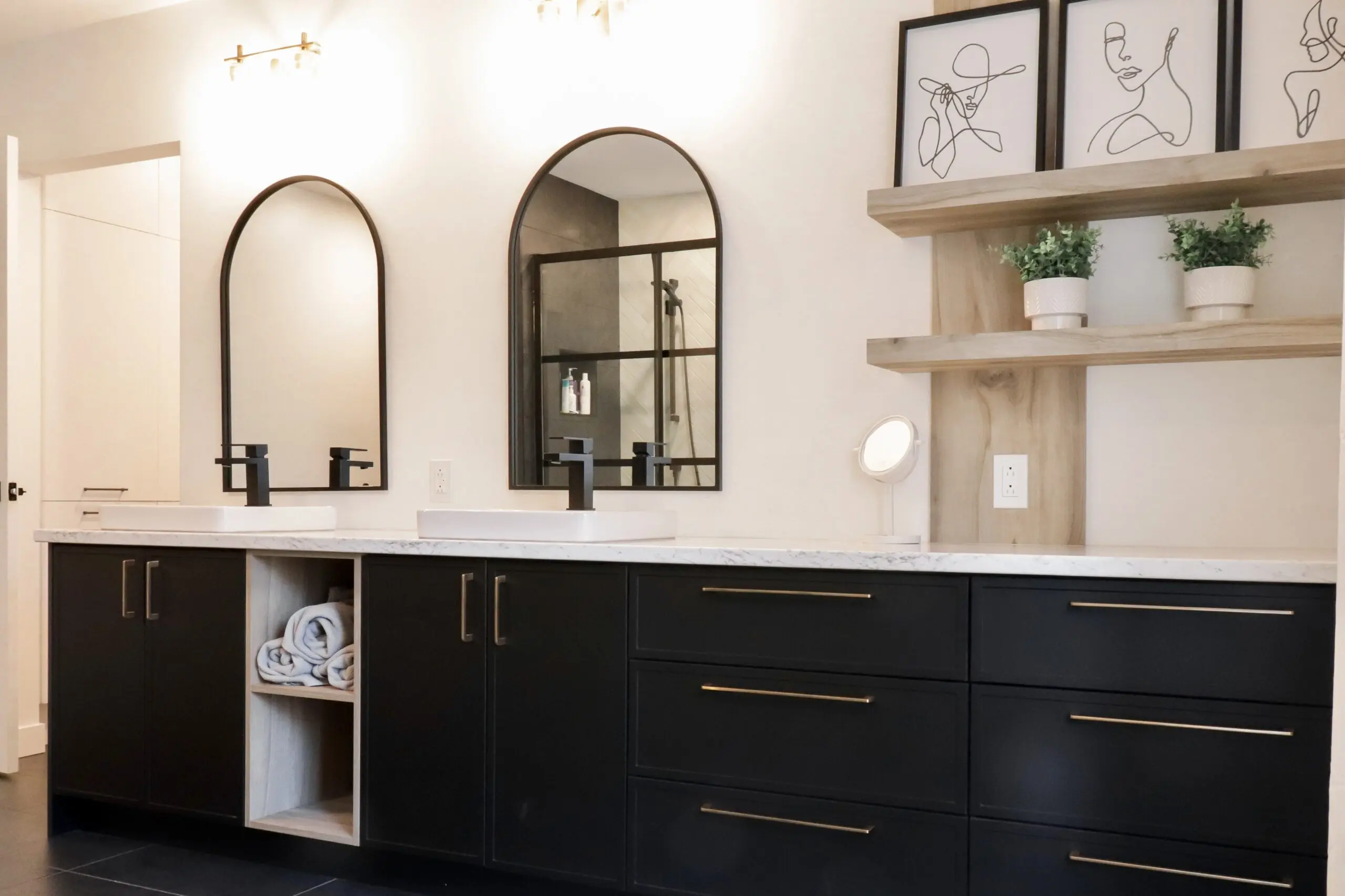 Salle de bains moderne avec double lavabo, armoires noires avec poignées dorées, deux miroirs cintrés, art minimaliste au mur, étagères avec plantes en pot, et serviettes soigneusement enroulées dans une étagère ouverte.