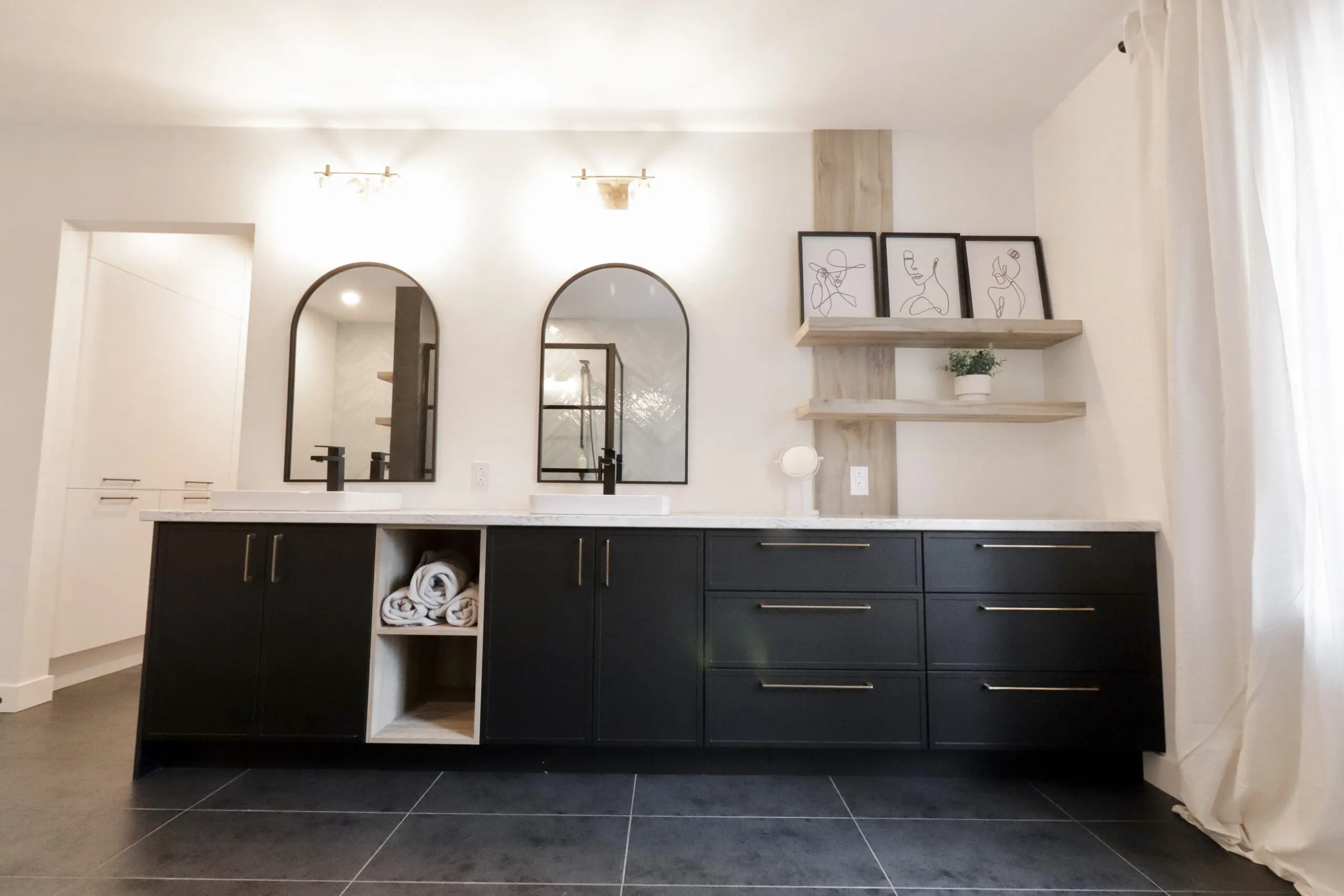 Salle de bains moderne avec double lavabo, armoires noires, poignées dorées, deux miroirs ovales, deux appliques murales, des étagères ouvertes avec des tableaux encadrés et une plante, des serviettes roulées dans un casier, et de grandes fenêtres avec des rideaux blancs.