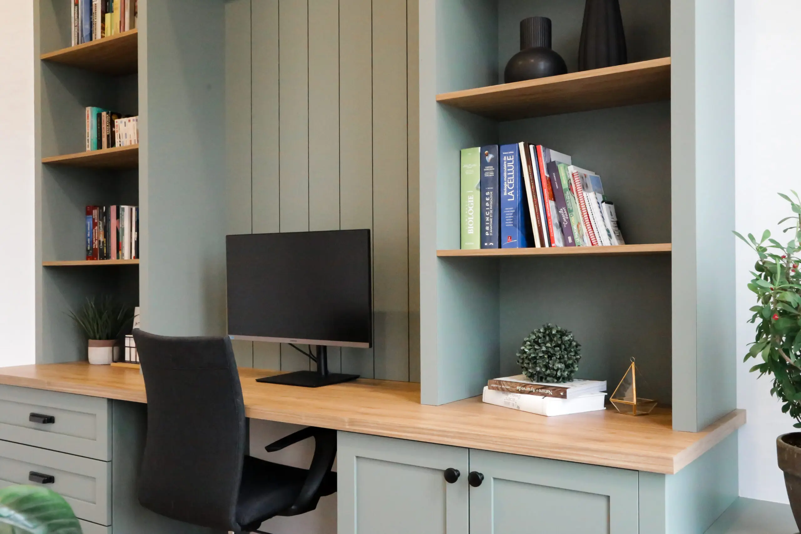 Un bureau à domicile moderne avec un bureau en bois, un écran d'ordinateur, des étagères remplies de livres et de décorations, une chaise de bureau noire et des plantes en pot. Les étagères et les armoires sont peintes en vert clair.