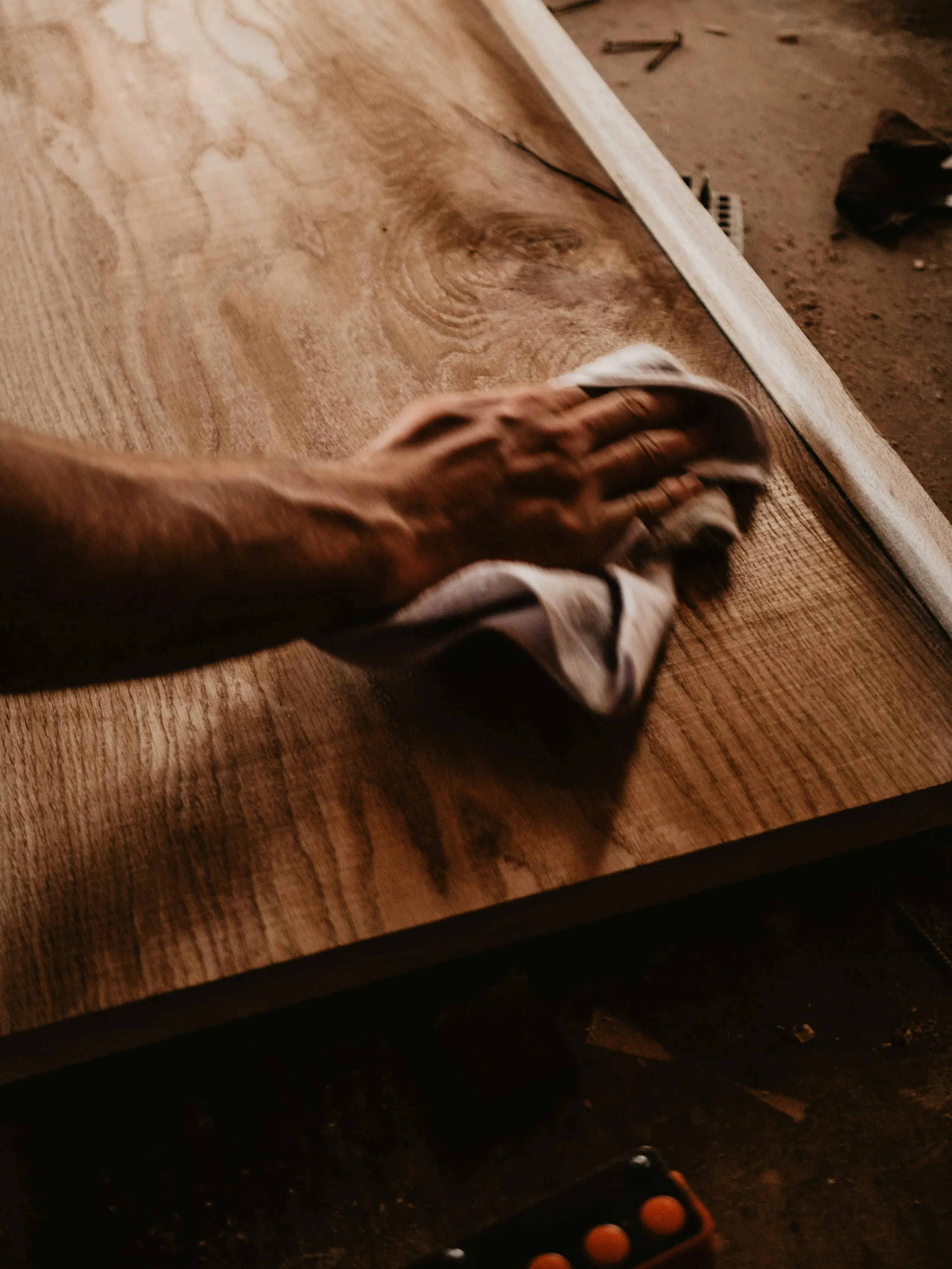La main d'une personne essuie ou polit une surface en bois à l'aide d'un chiffon, donnant au bois une finition propre et lisse. Cette scène évoque le travail du bois ou l'entretien de meubles dans un atelier.