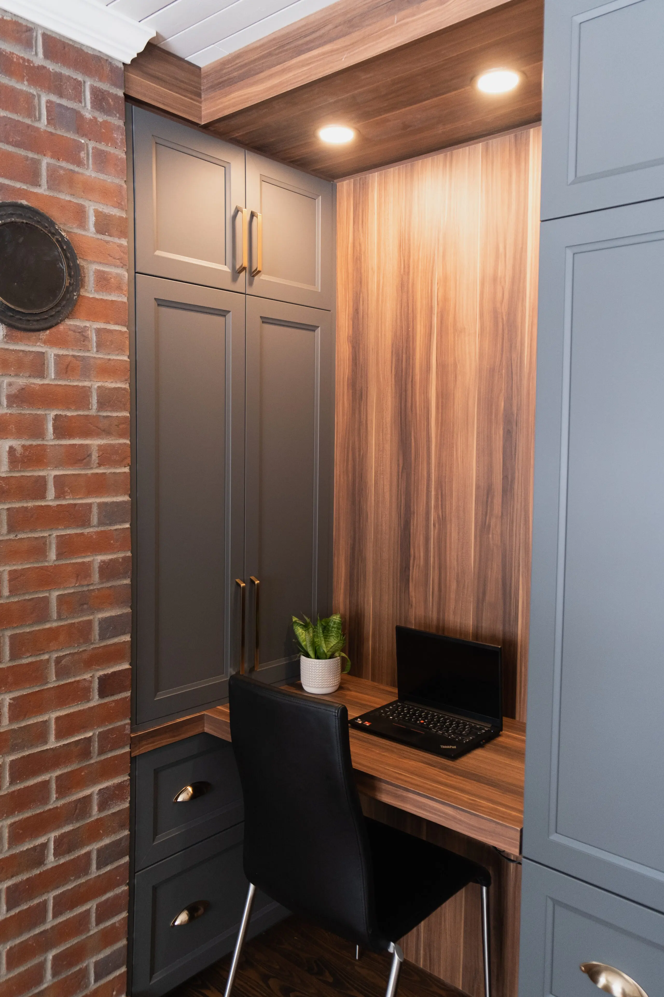 Un petit bureau à domicile avec des armoires grises, un bureau en bois, une chaise noire, un ordinateur portable fermé, une petite plante en pot et un éclairage encastré au plafond, le tout contre un mur de briques.