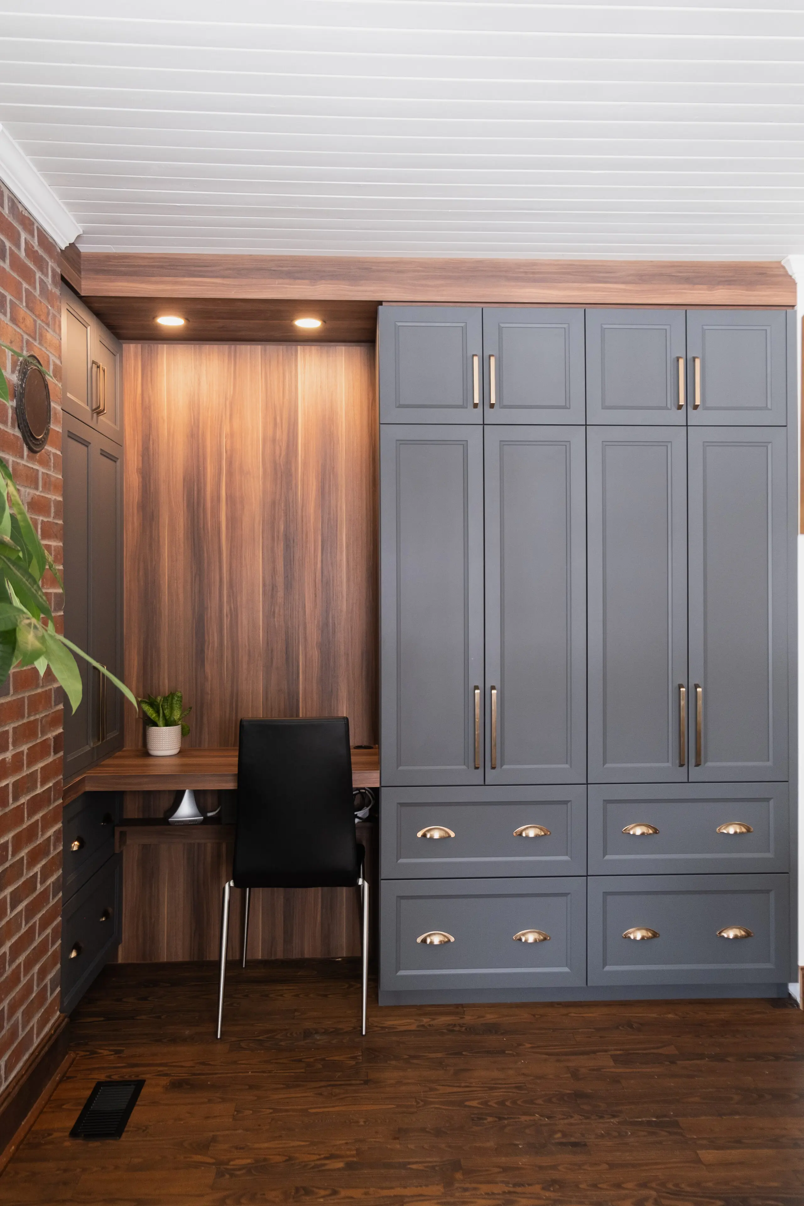 Un espace de travail moderne intégré avec des armoires grises, des panneaux de bois, des plafonniers, une chaise noire et une petite plante en pot sur le bureau. Un mur de briques et une partie d'une plante à feuilles vertes sont visibles sur la gauche.