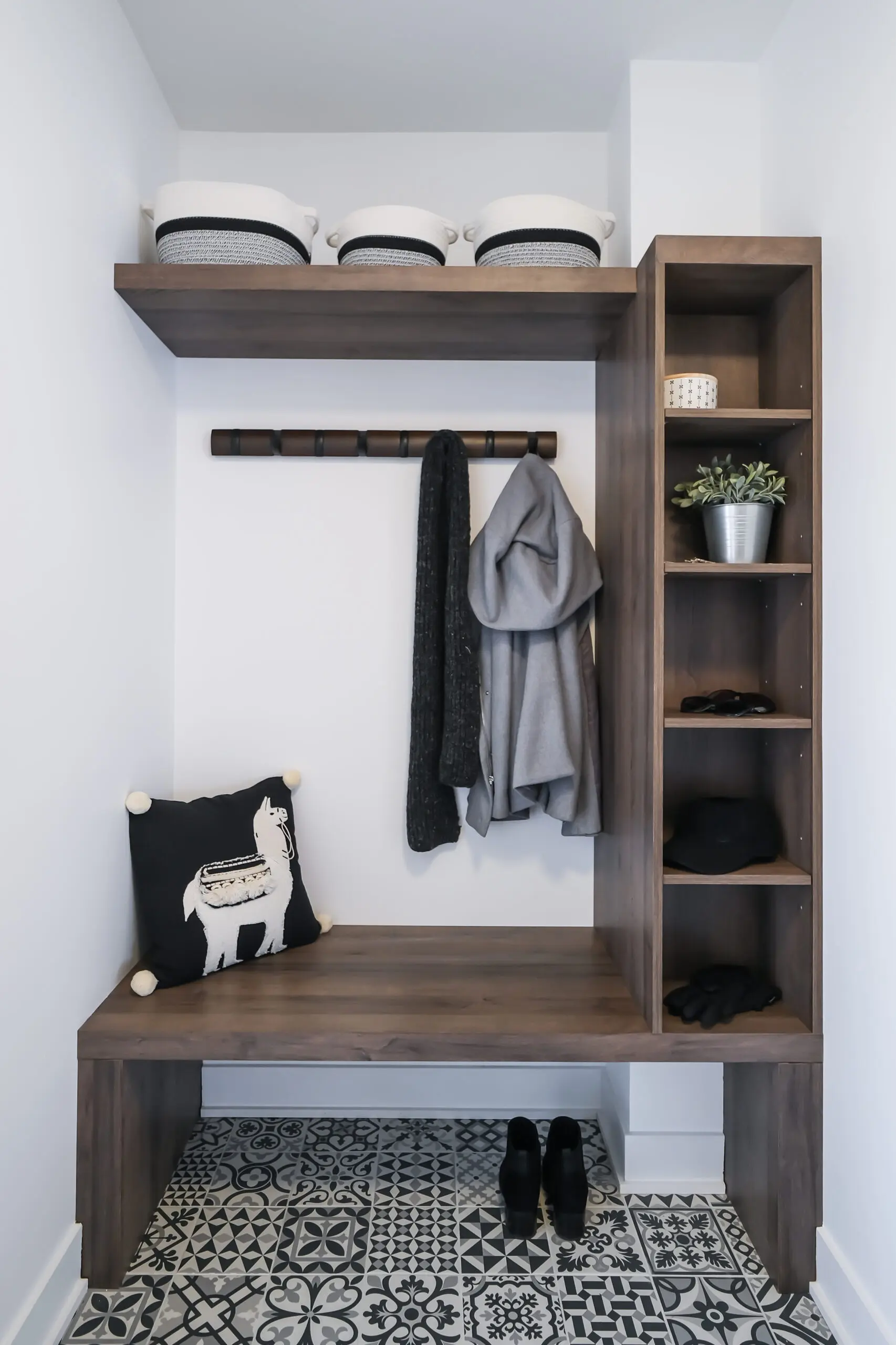 Un vestiaire moderne avec un sol en carrelage à motifs, un banc en bois, des étagères, des paniers de rangement, une plante, des patères avec des vestes, un coussin avec un motif de lama et une paire de chaussures noires sur le sol.