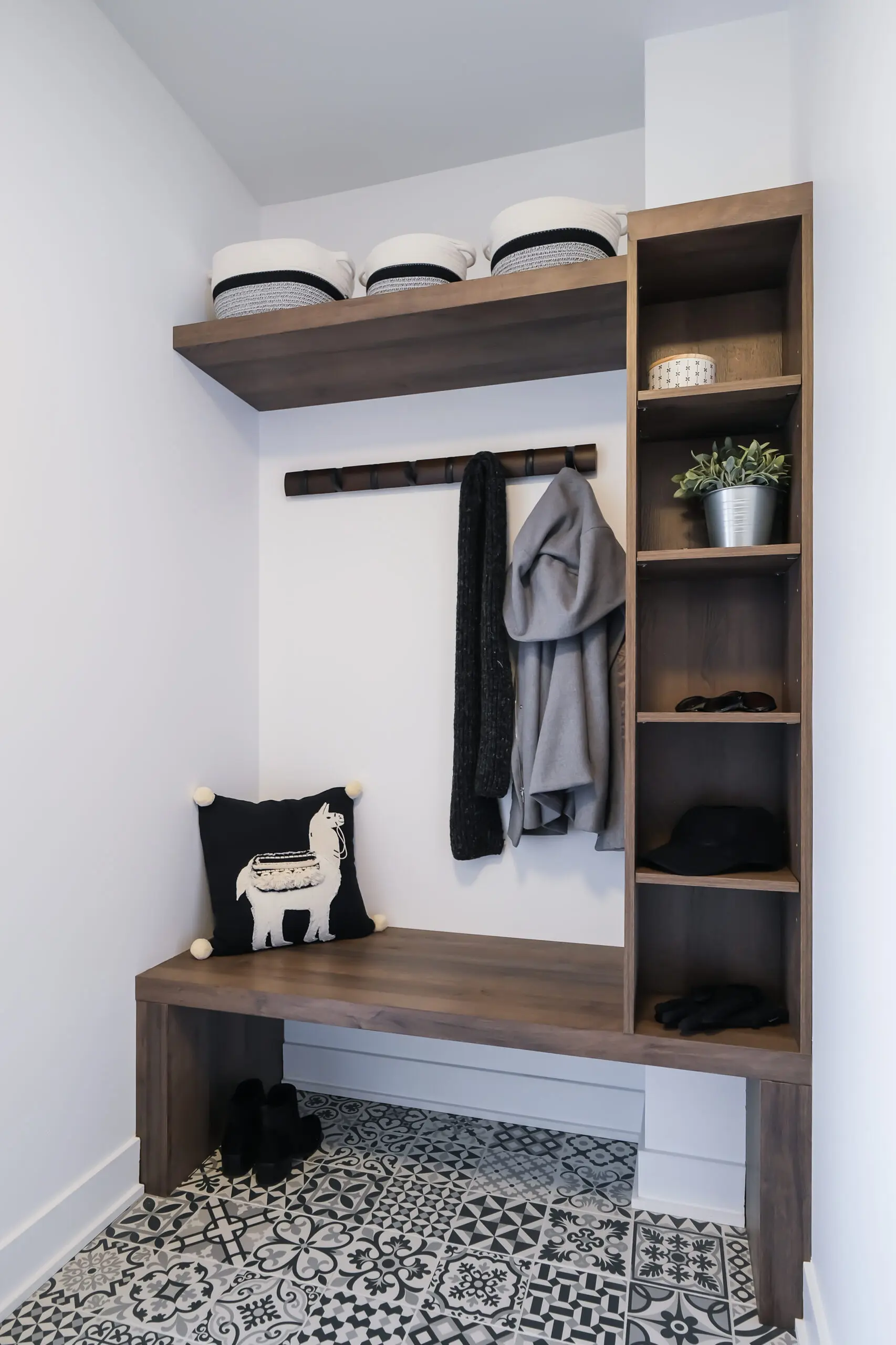 Une petite entrée présente un carrelage à motifs, un banc en bois avec un oreiller en forme de lama, des étagères ouvertes avec des paniers, des patères avec des vestes, une plante en pot, des chapeaux et des chaussures bien rangées.