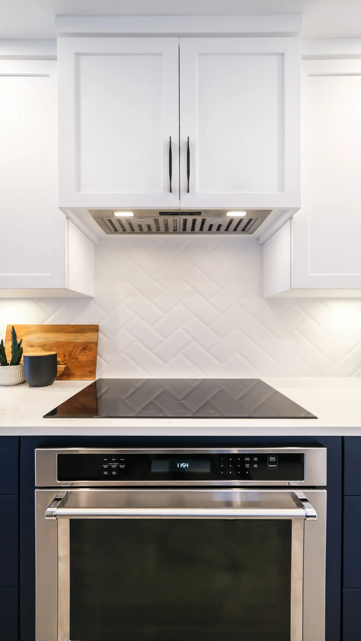 Cuisine moderne avec un four en acier inoxydable, une table de cuisson en verre noir, des armoires blanches et un dosseret en carreaux blancs à chevrons ; une petite plante et une planche à découper se trouvent sur le comptoir.