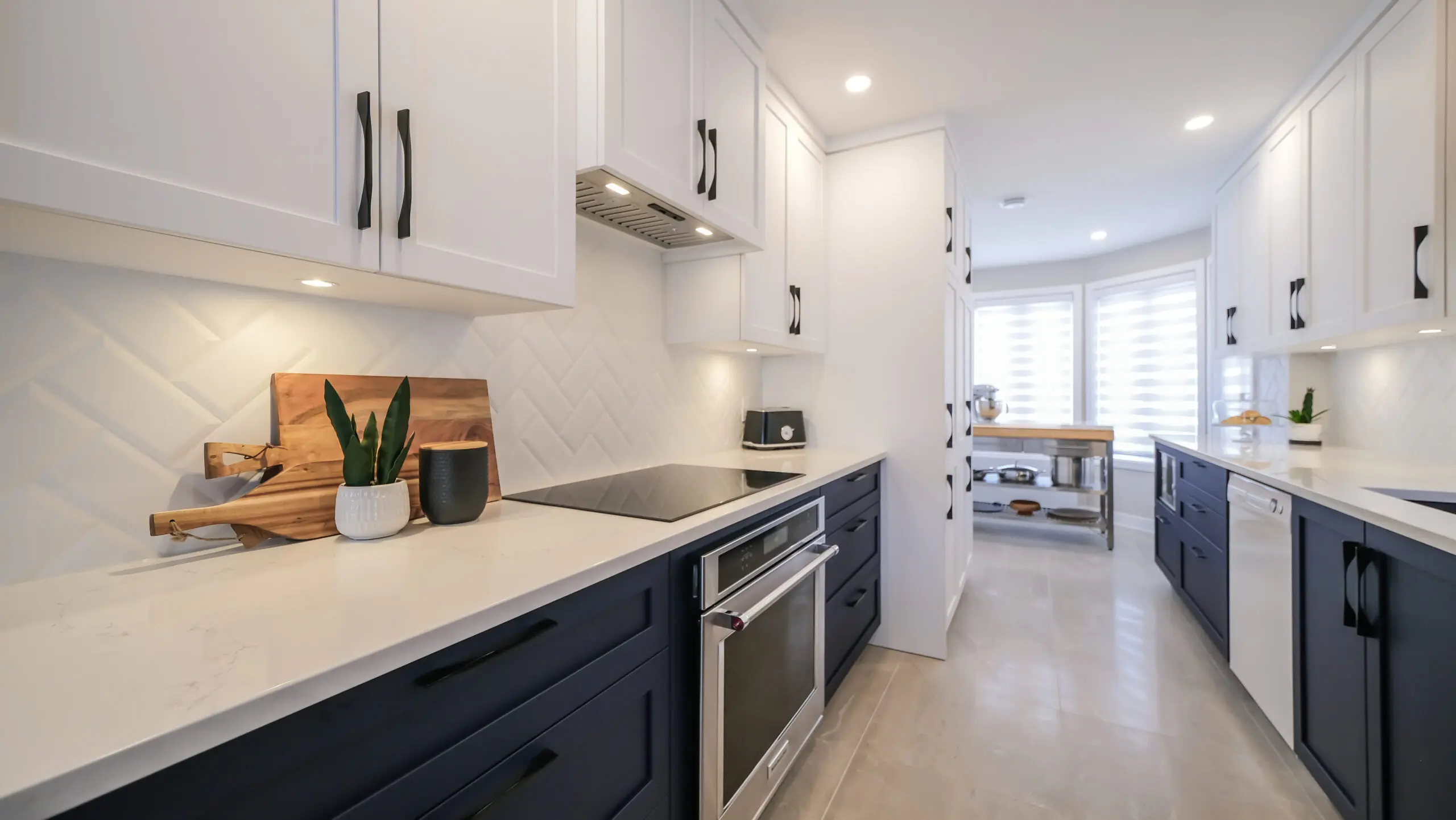 Cuisine moderne avec armoires supérieures blanches, armoires inférieures foncées, comptoirs blancs, cuisinière encastrée, éclairage sous l'armoire et objets décoratifs, menant à un coin repas lumineux avec de grandes fenêtres.