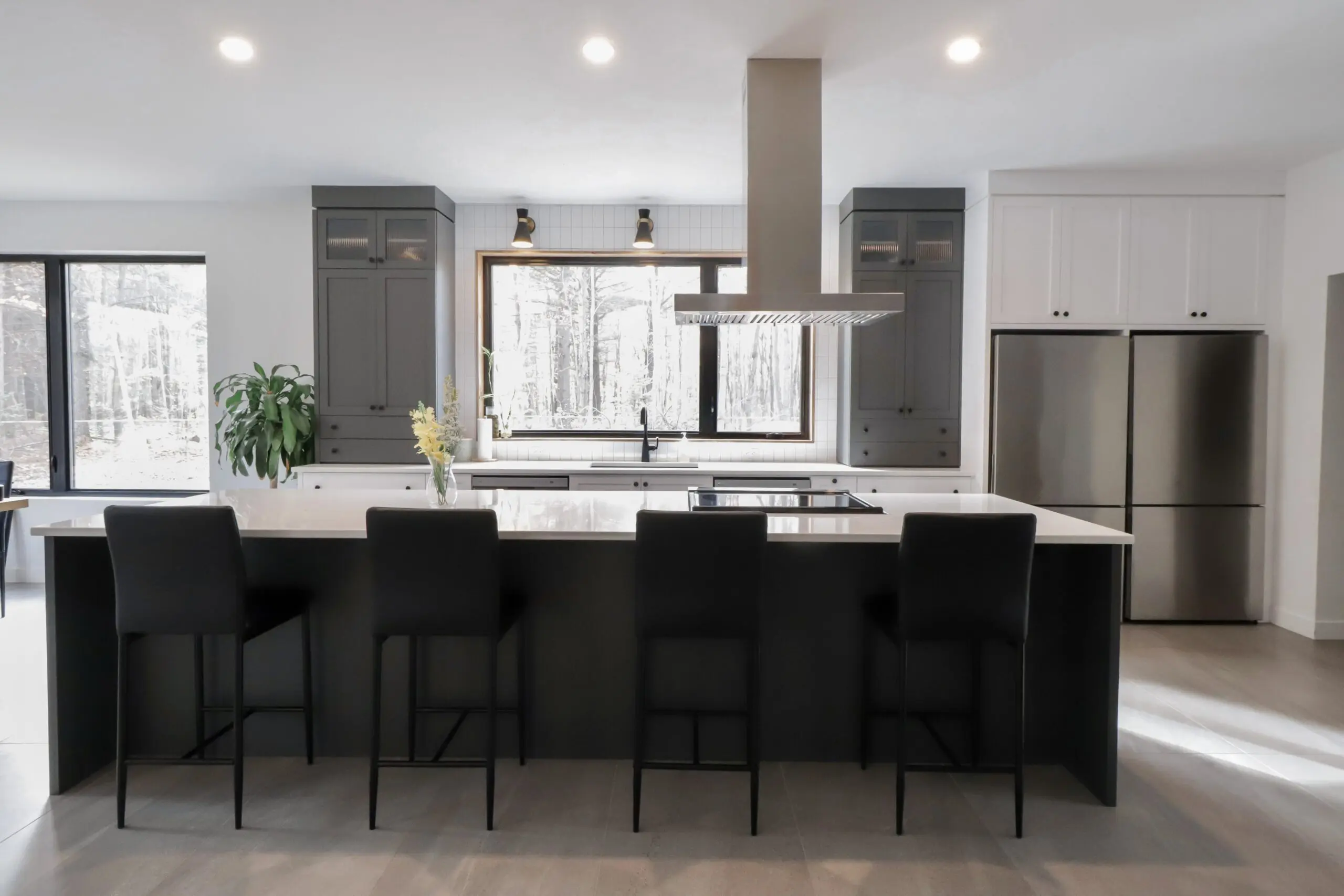 Cuisine moderne avec un grand îlot, quatre chaises noires, des appareils en acier inoxydable, des armoires sombres, une grande fenêtre, une plante et un décor minimaliste. La lumière naturelle pénètre dans la pièce par la fenêtre.