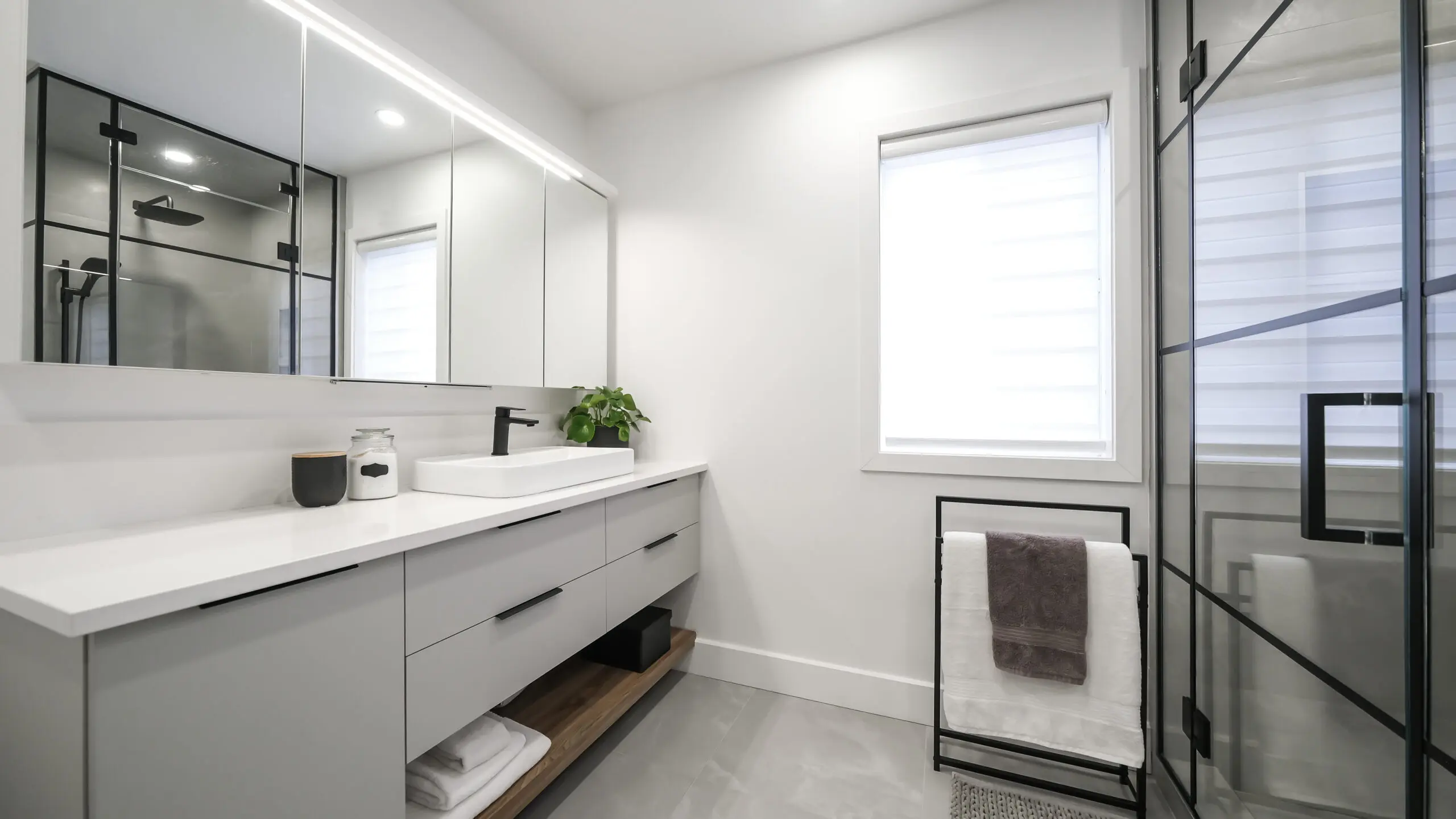 Salle de bains moderne avec murs blancs, grand miroir, comptoir blanc épuré, lavabo avec robinet noir, cabine de douche en verre, et serviettes pliées sur un porte-serviettes noir près d'une fenêtre. Une petite plante orne le comptoir.