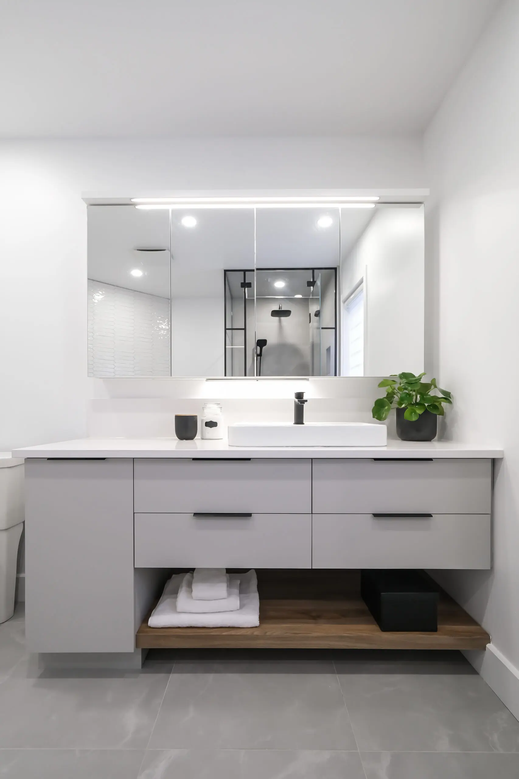 Salle de bains moderne avec un grand miroir, un meuble-lavabo gris, un comptoir blanc, un lavabo, un robinet noir, une petite plante, des serviettes soigneusement pliées sur une étagère en bois et une cabine de douche en verre qui se reflète dans le miroir.